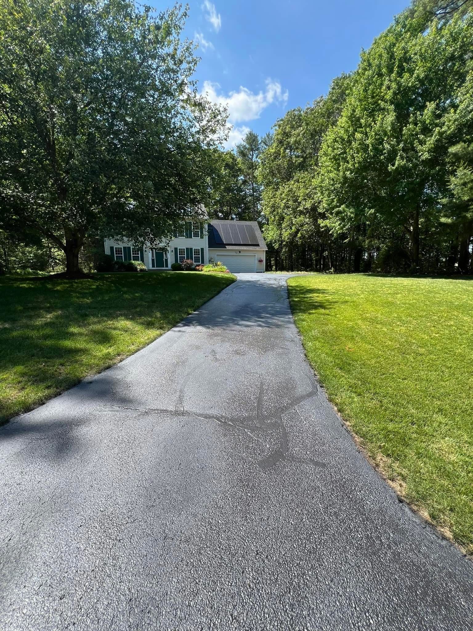 A paved driveway leads to a two-story house surrounded by mature trees and lush green lawns under a blue sky.