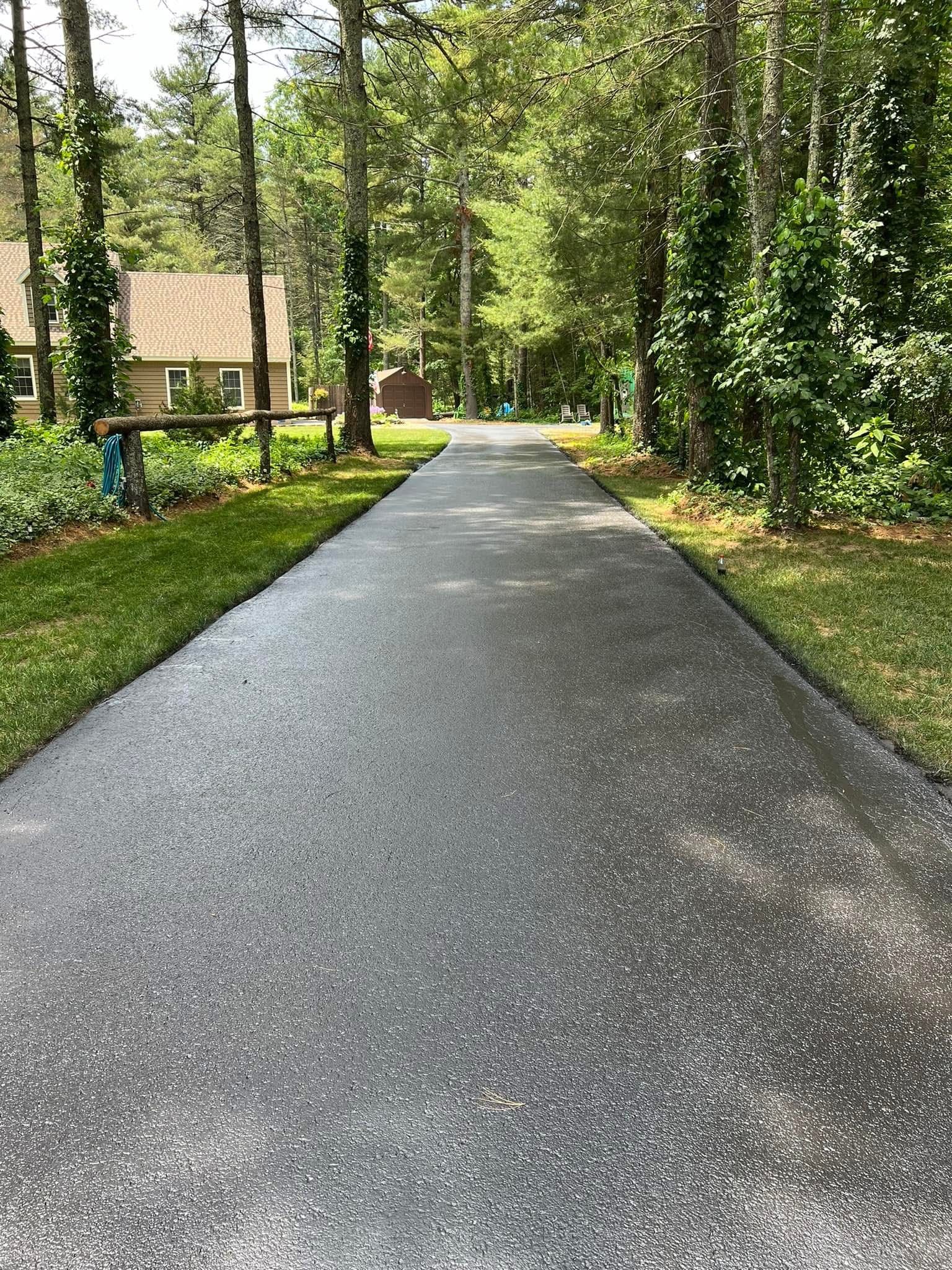 A long, freshly paved asphalt driveway stretches through a wooded area toward a house in the distance.