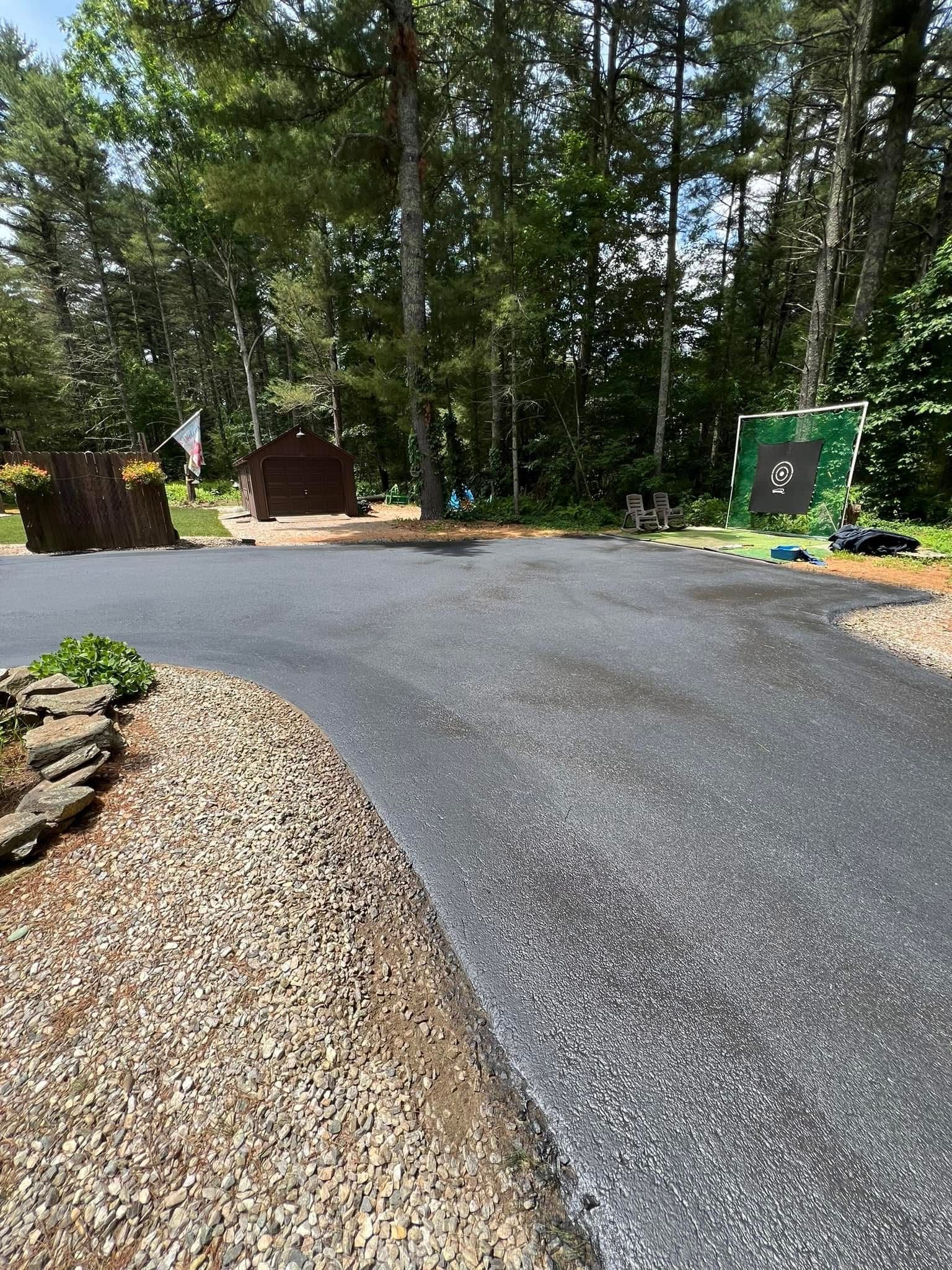 A newly paved asphalt driveway curves through a wooded area toward a brown shed and a green target stand.