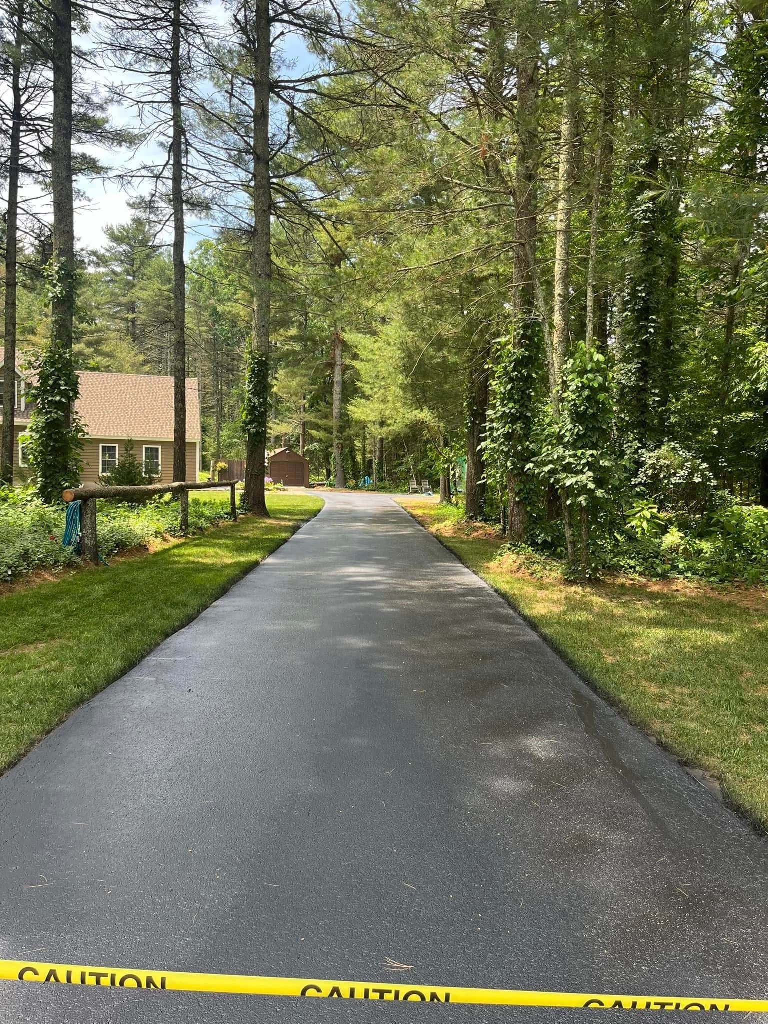 A freshly paved driveway leads through a wooded area toward a house, blocked by yellow caution tape in the foreground.