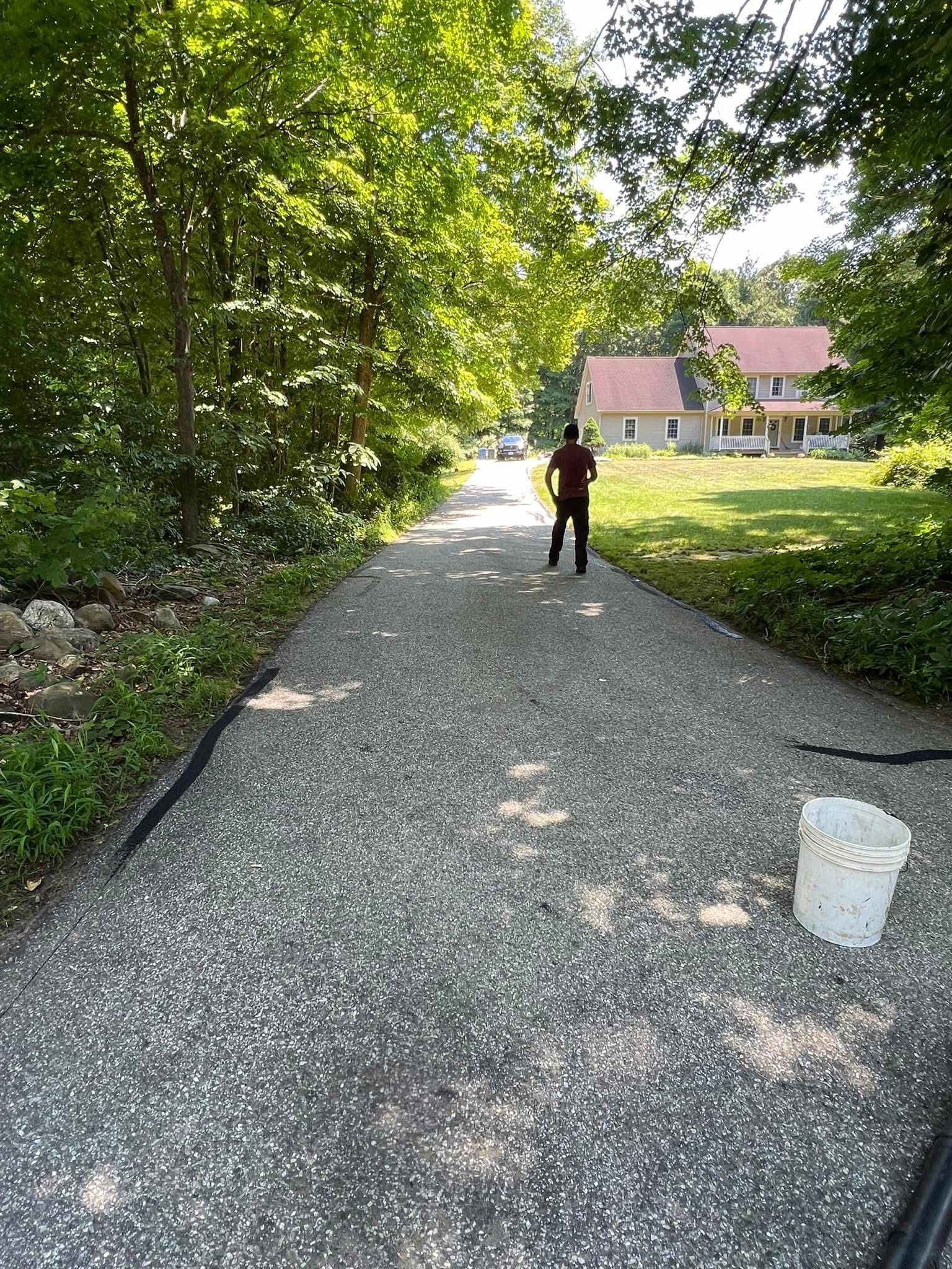 A person walks up a gravel driveway lined by trees, leading toward a house on a sunny day with a bucket in the foreground.