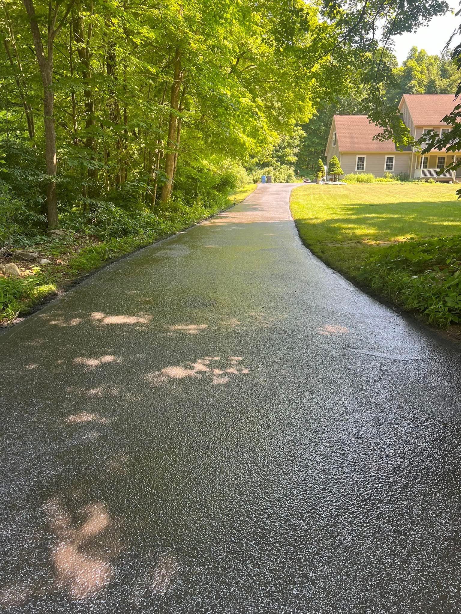 A glistening, freshly sealed dark asphalt driveway curves past a grassy lawn and lush green trees towards a house.