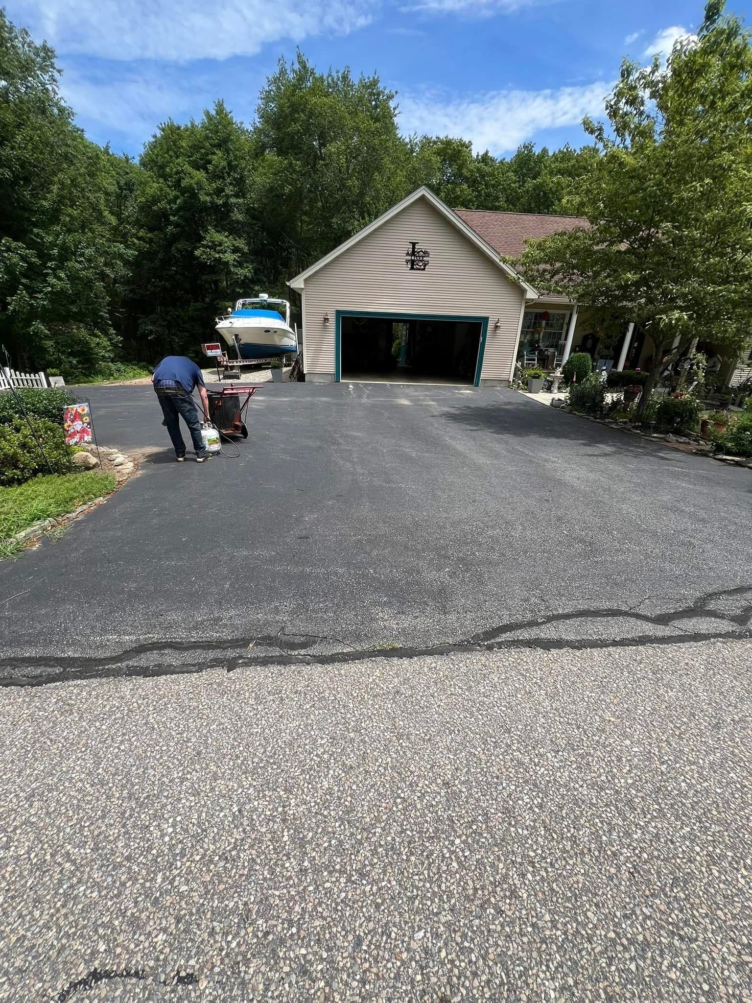 A worker applies sealant to a driveway in front of a garage and house with a boat parked nearby.