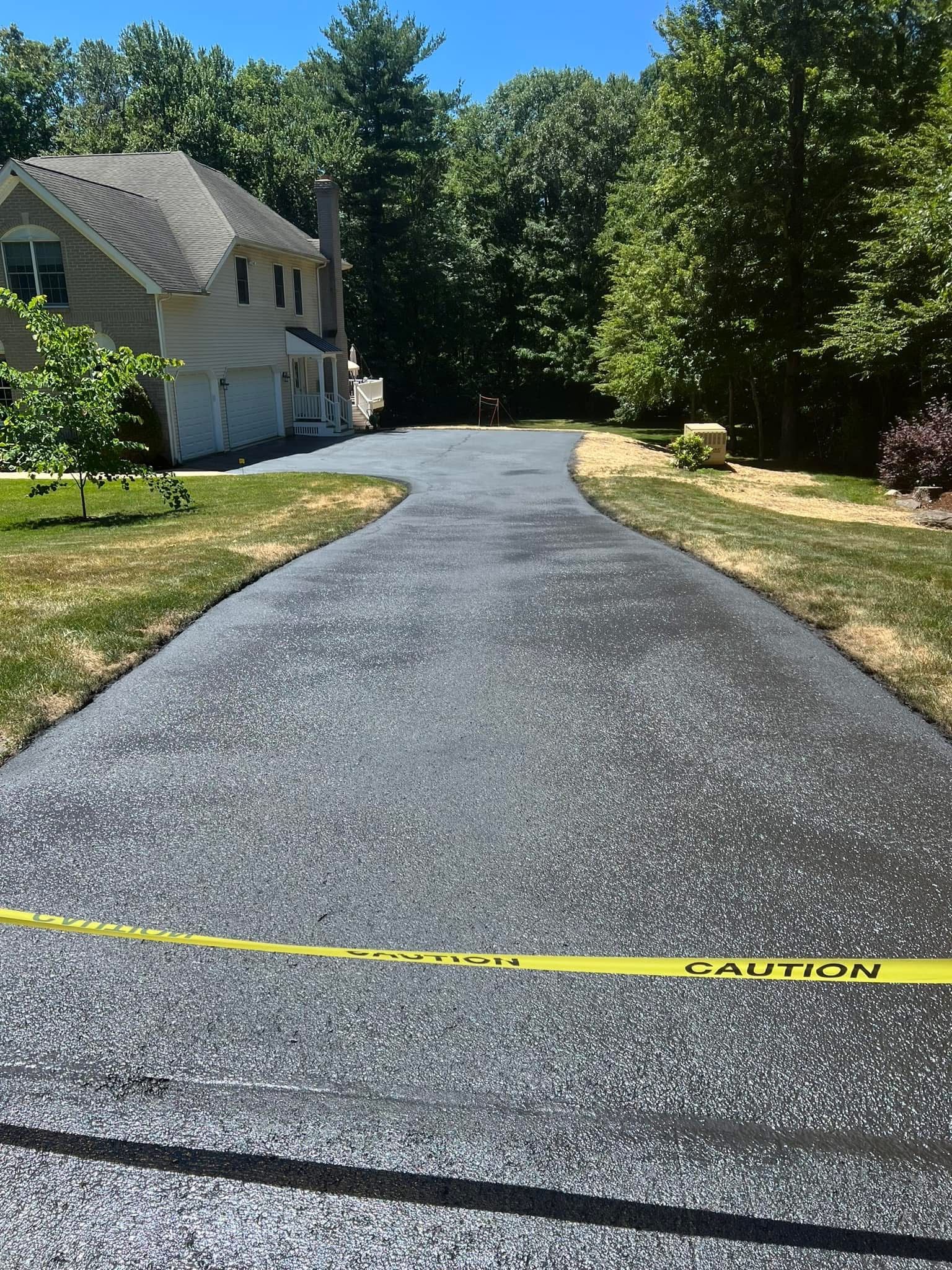 A freshly paved asphalt driveway leading toward a two-story house, blocked by yellow caution tape.