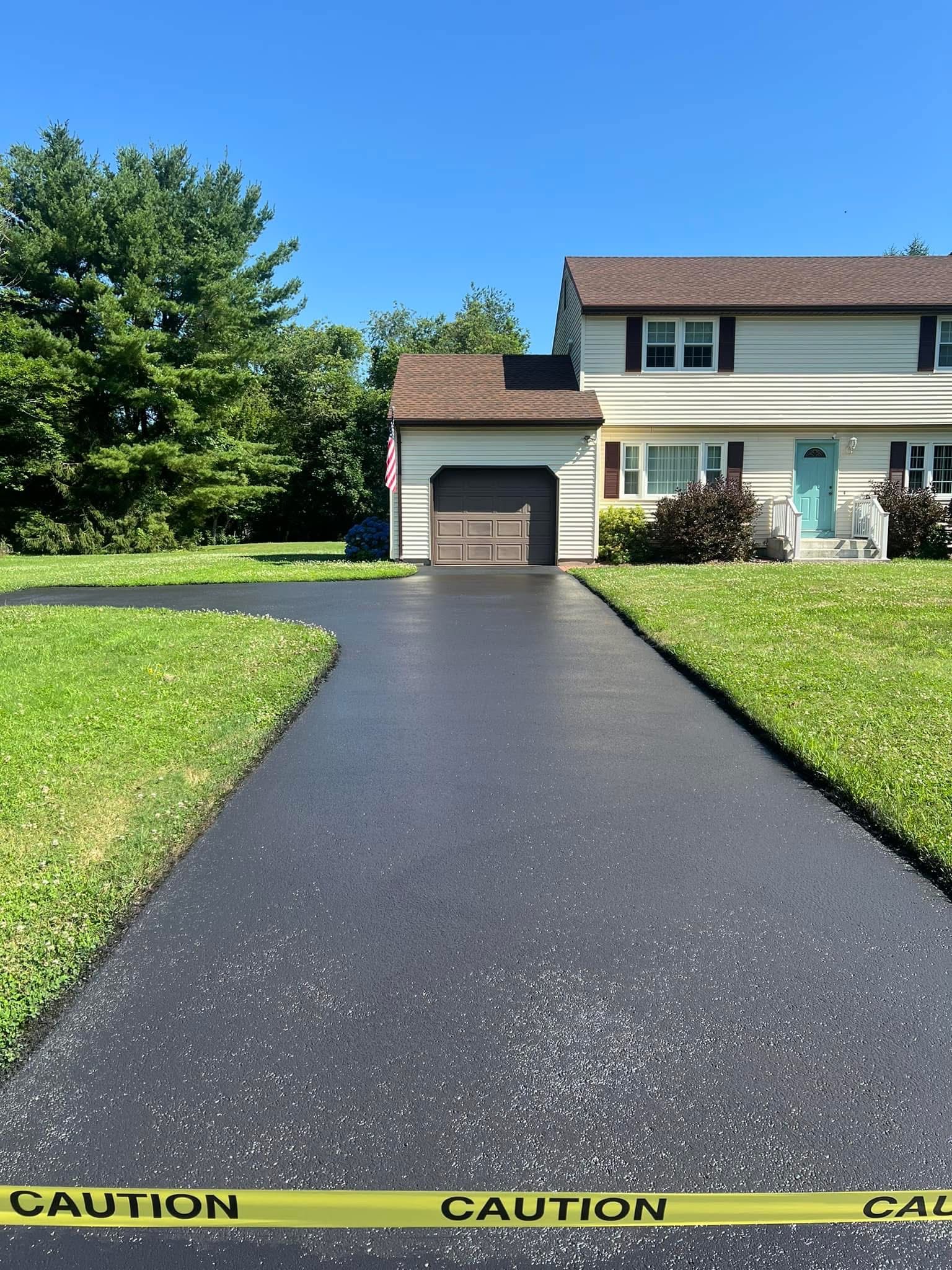 A freshly paved, black driveway leads to a suburban house with a garage, blocked off by yellow caution tape.