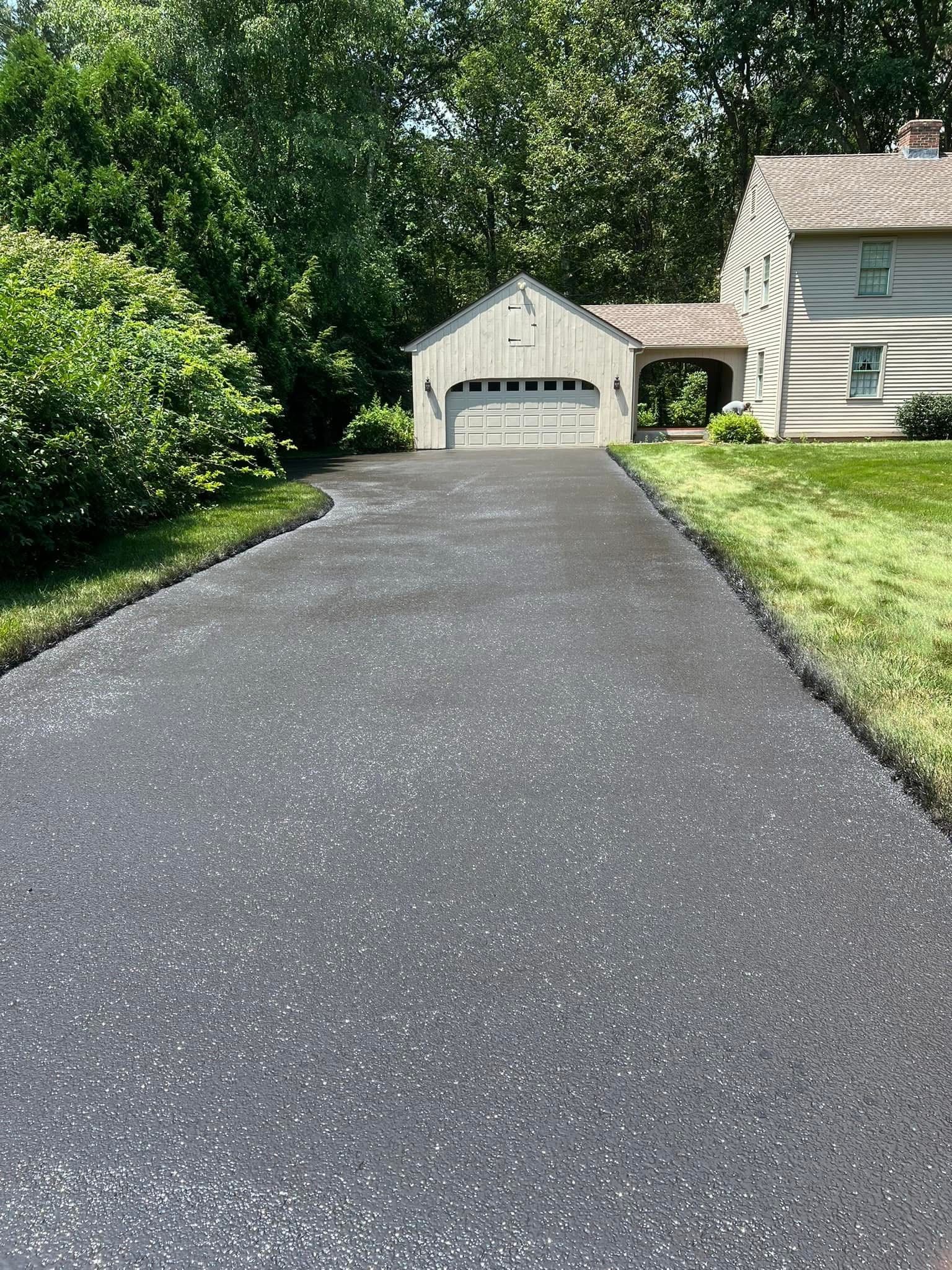 A paved driveway leading toward a detached garage and house nestled among lush green trees and lawn.