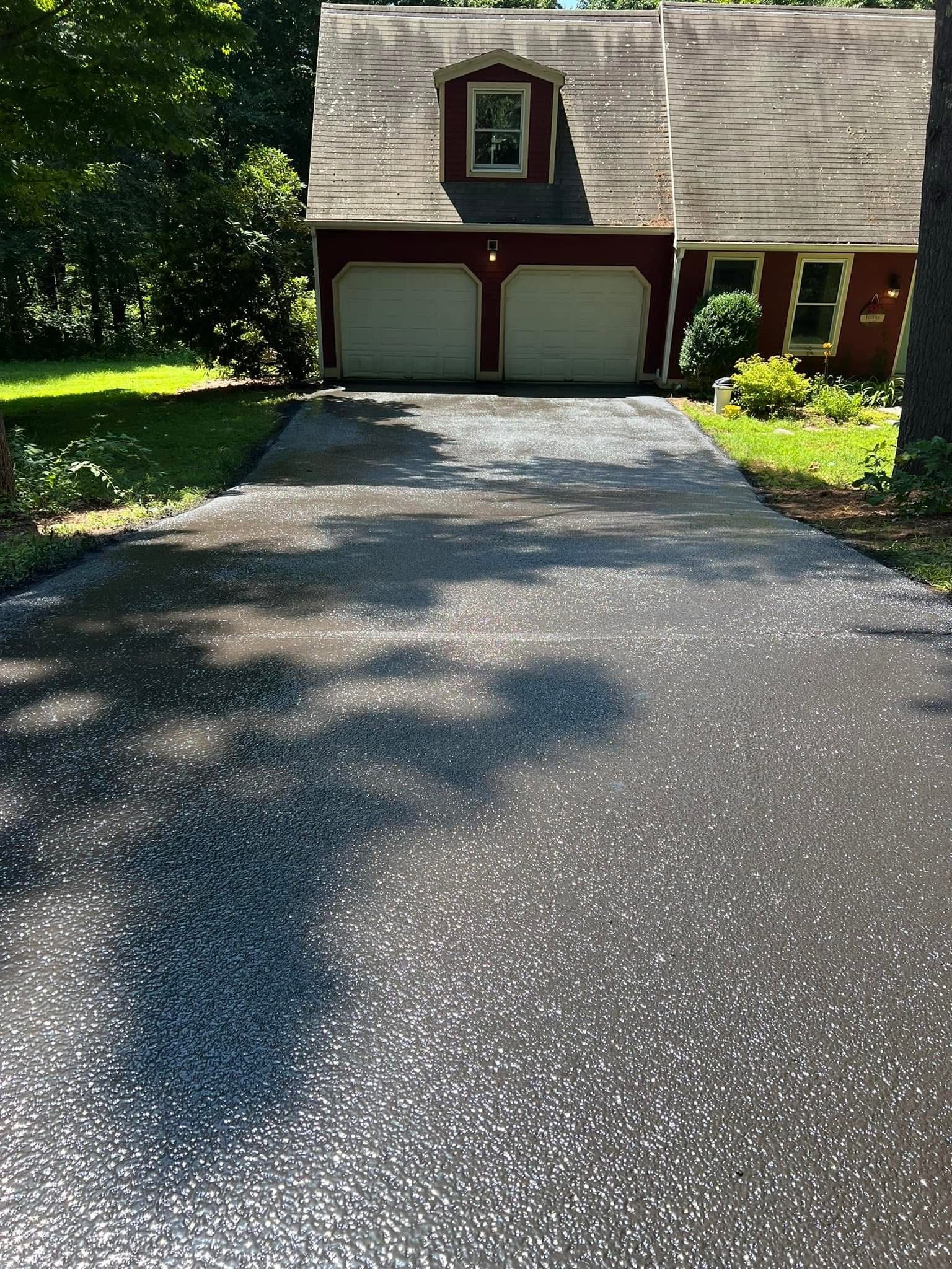 A black, freshly paved residential driveway leads to a house with a red garage and a dormer window.