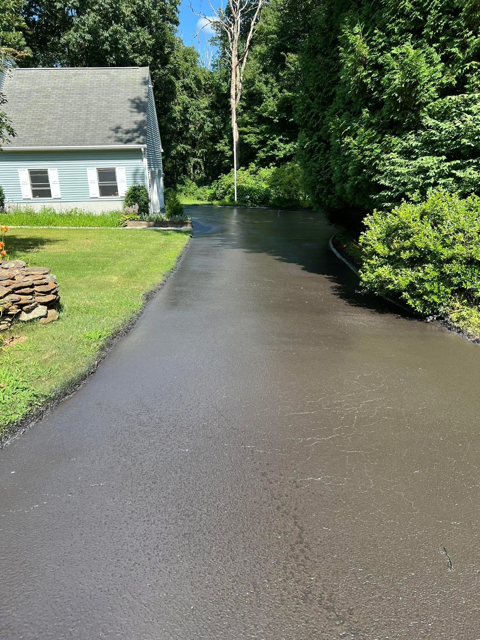 A newly paved dark asphalt driveway leads toward a light blue house surrounded by green trees and a grassy lawn.