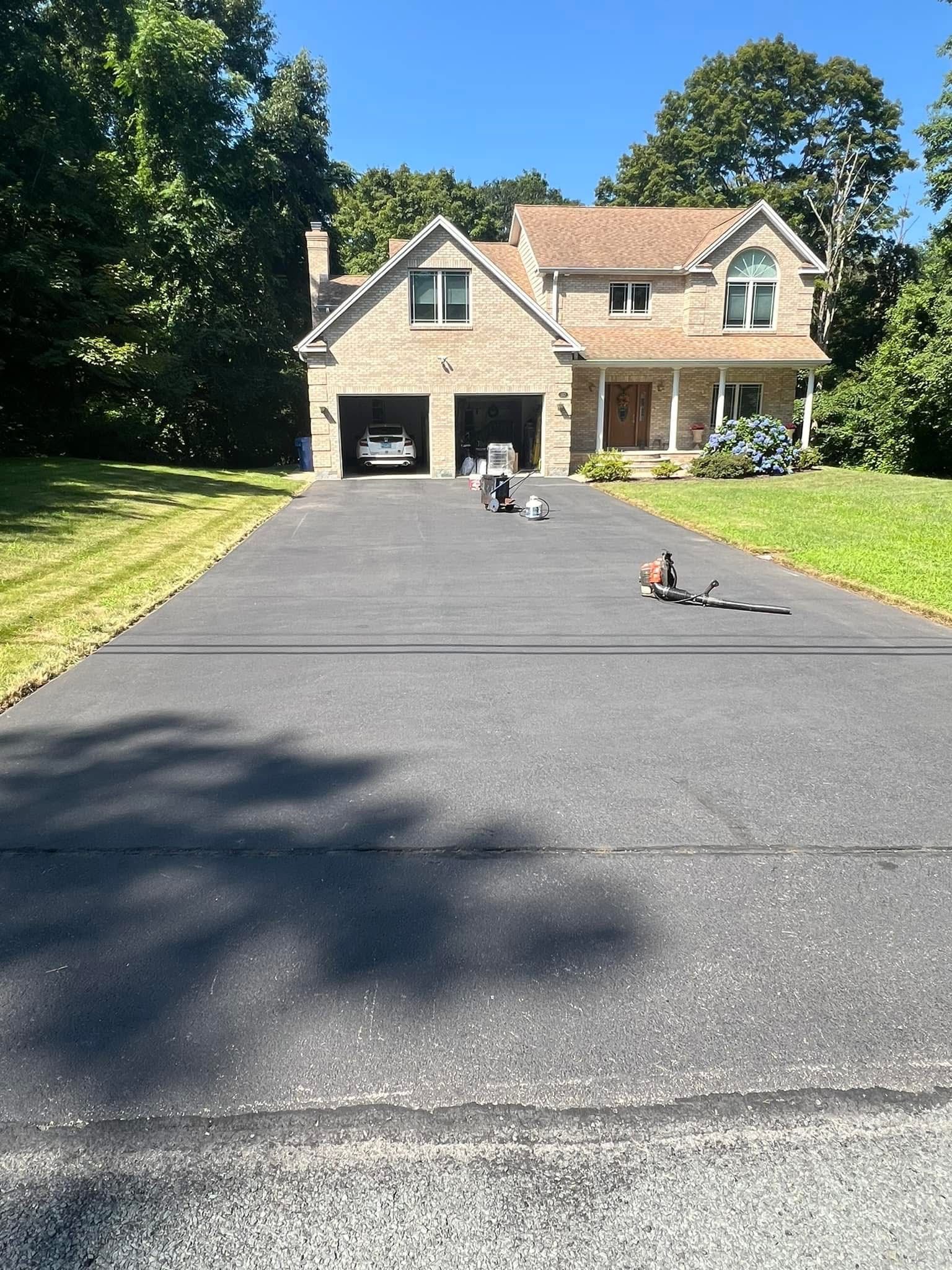 A freshly paved asphalt driveway leading to a two-story brick house with a two-car garage on a sunny day.