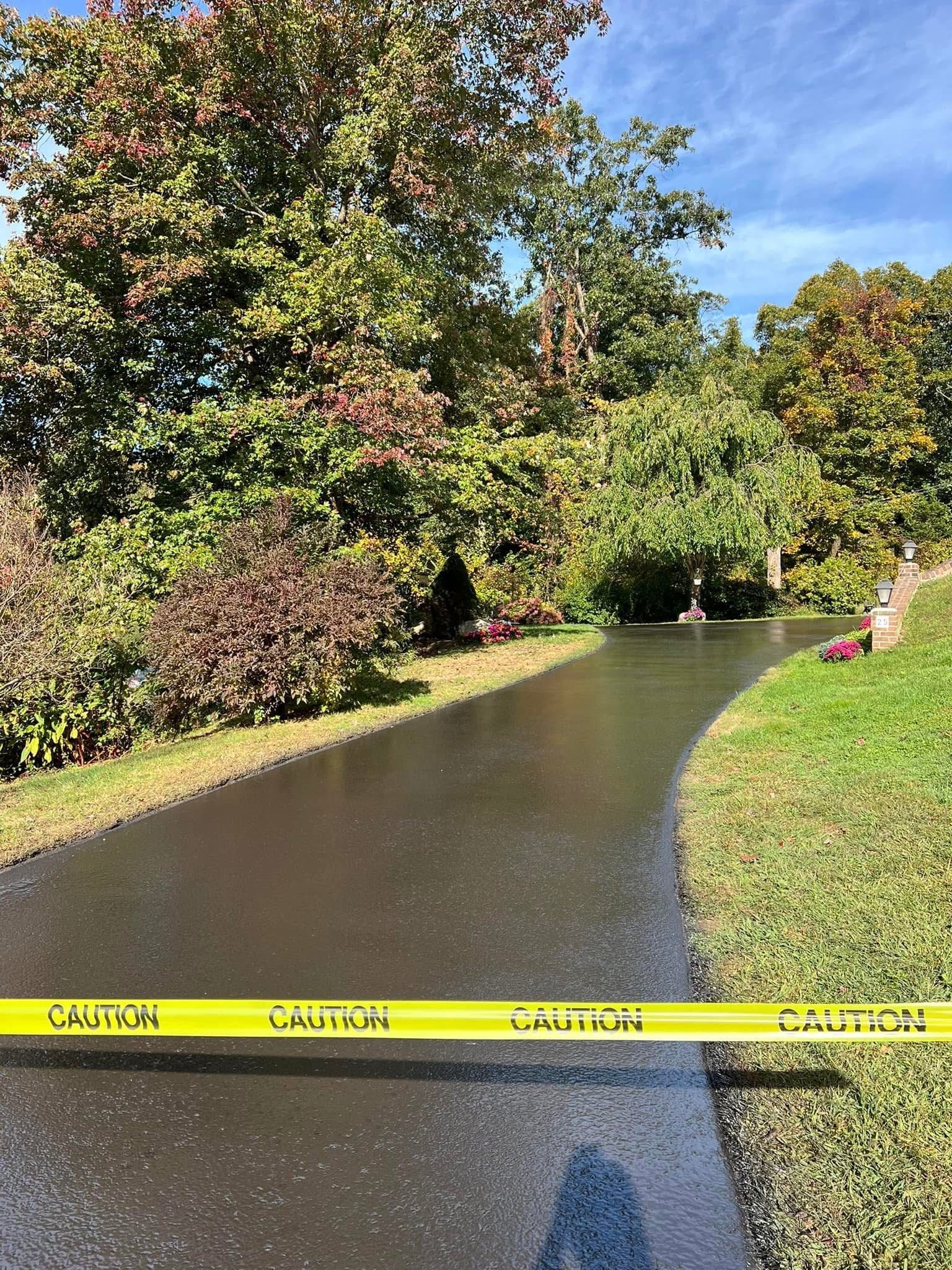 A freshly paved, dark asphalt driveway lined with trees and grass, blocked by a yellow caution tape.