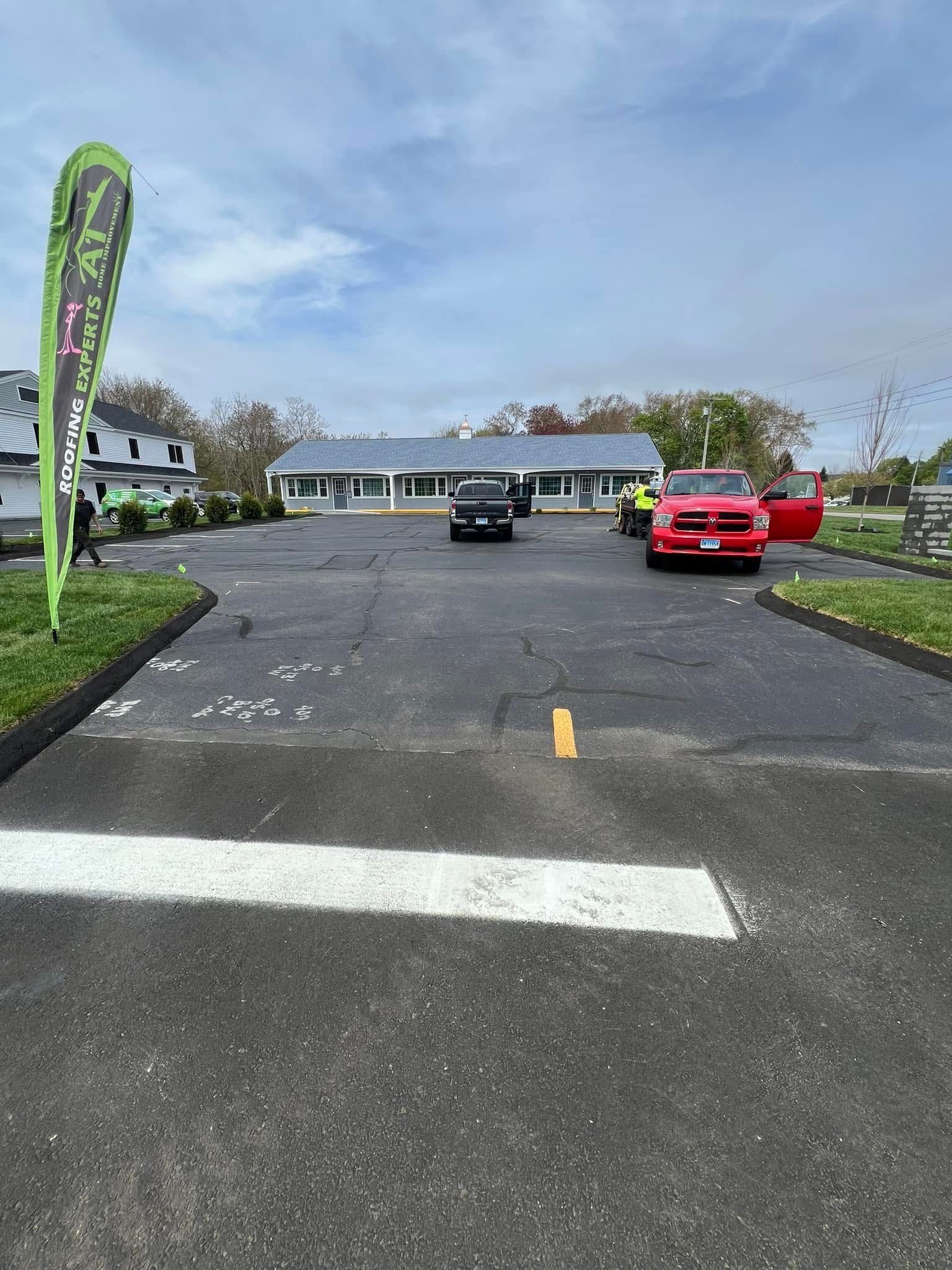 A parking lot with a red truck, a dark car, and a green promotional flag in front of a long, low building under a blue sky.