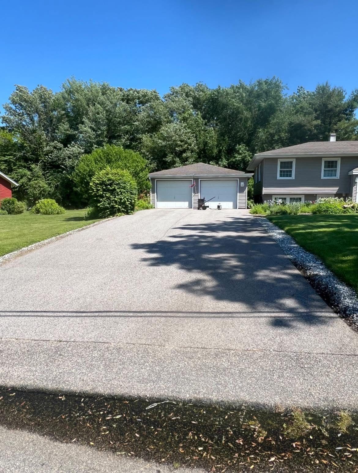 A paved driveway leads to a two-car garage next to a gray house under a clear blue sky, surrounded by green trees and grass.