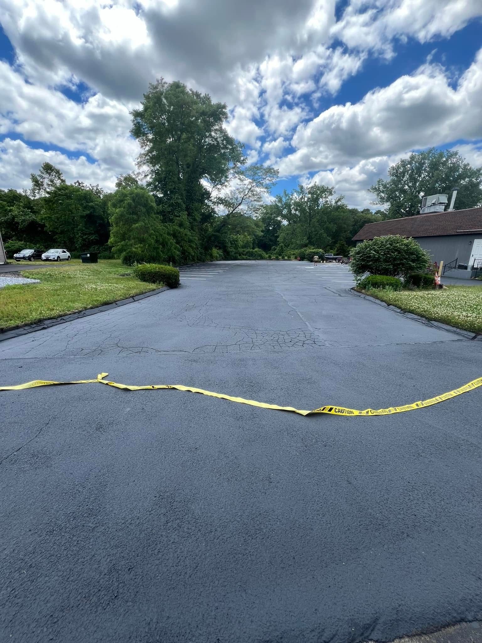 A freshly paved asphalt parking lot under a cloudy blue sky, marked off by a strip of yellow caution tape.