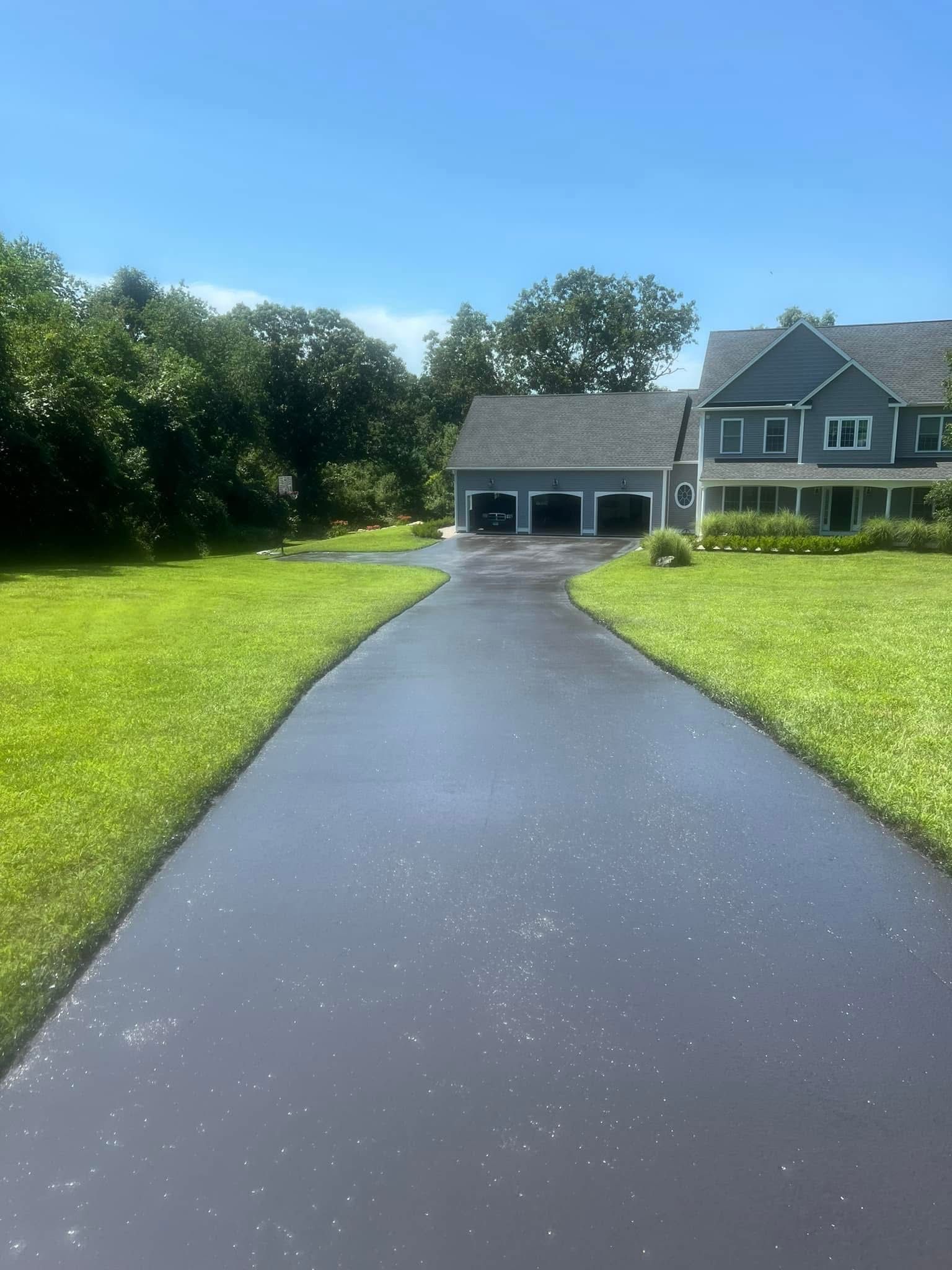 A freshly sealed black asphalt driveway leads toward a two-story gray house with a three-car garage and green lawn.