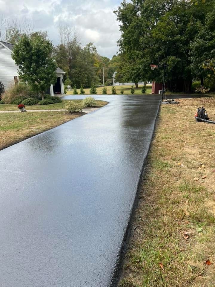A freshly sealed, wet asphalt driveway leading toward a house on a cloudy day, bordered by green lawn and trees.