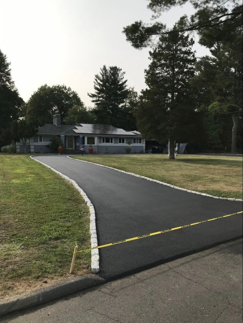 A freshly paved asphalt driveway with cobblestone edging leads to a single-story house surrounded by trees and lawn.