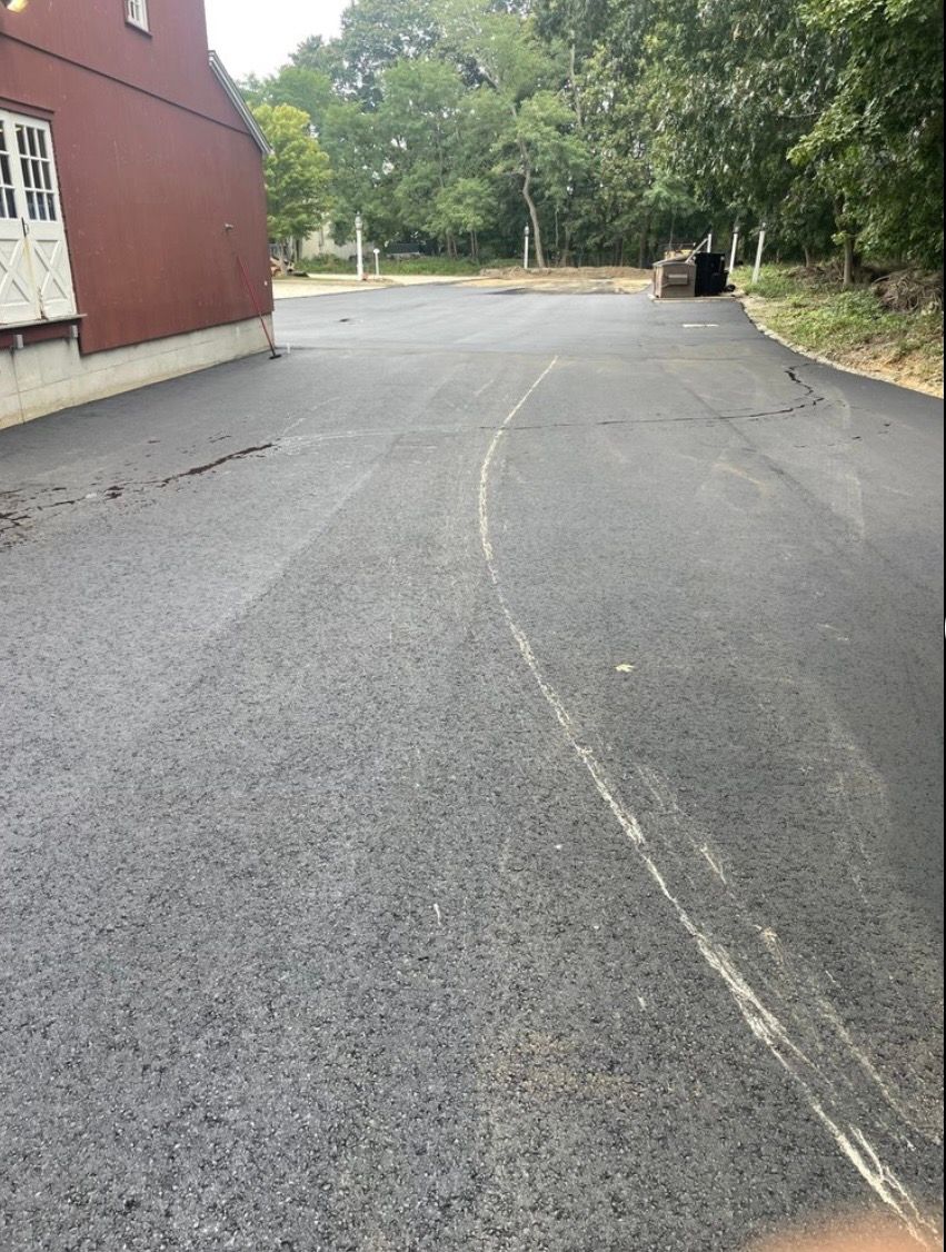 An asphalt driveway leads past a red building toward a wooded area under an overcast sky.