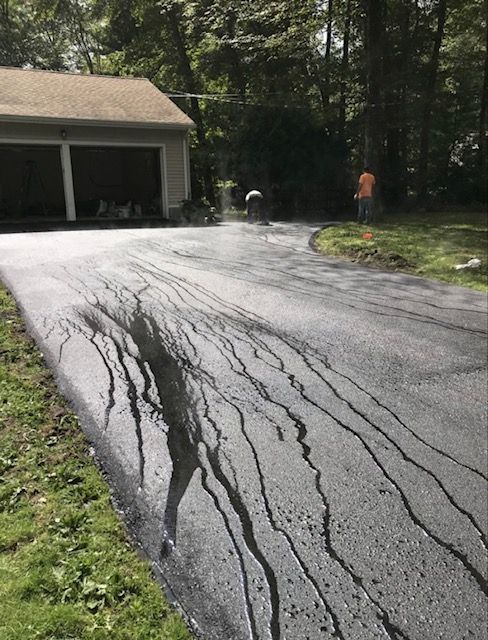 A driveway undergoing asphalt sealing, with dark liquid sealant running down the sloped surface toward the lawn.