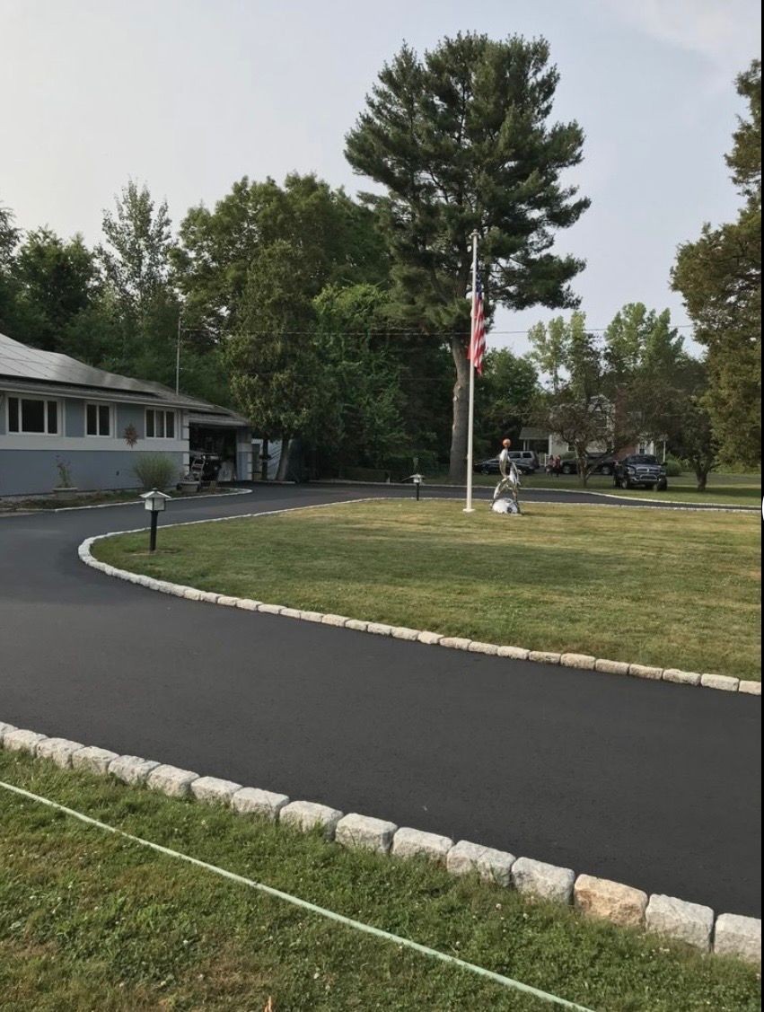 A curved asphalt driveway with stone edging leads to a building, with a flagpole on a grassy lawn under a hazy sky.