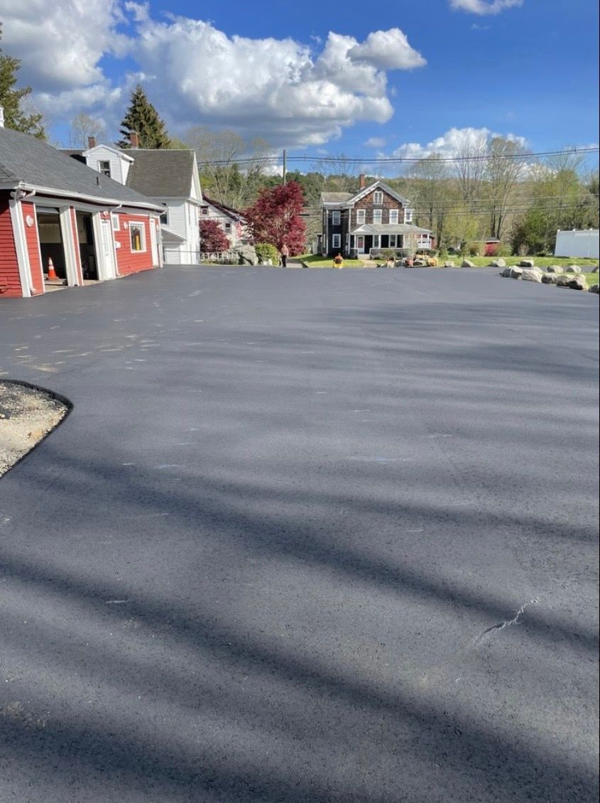 A newly paved asphalt driveway in front of a red building with a white house and trees in the background under a blue sky.