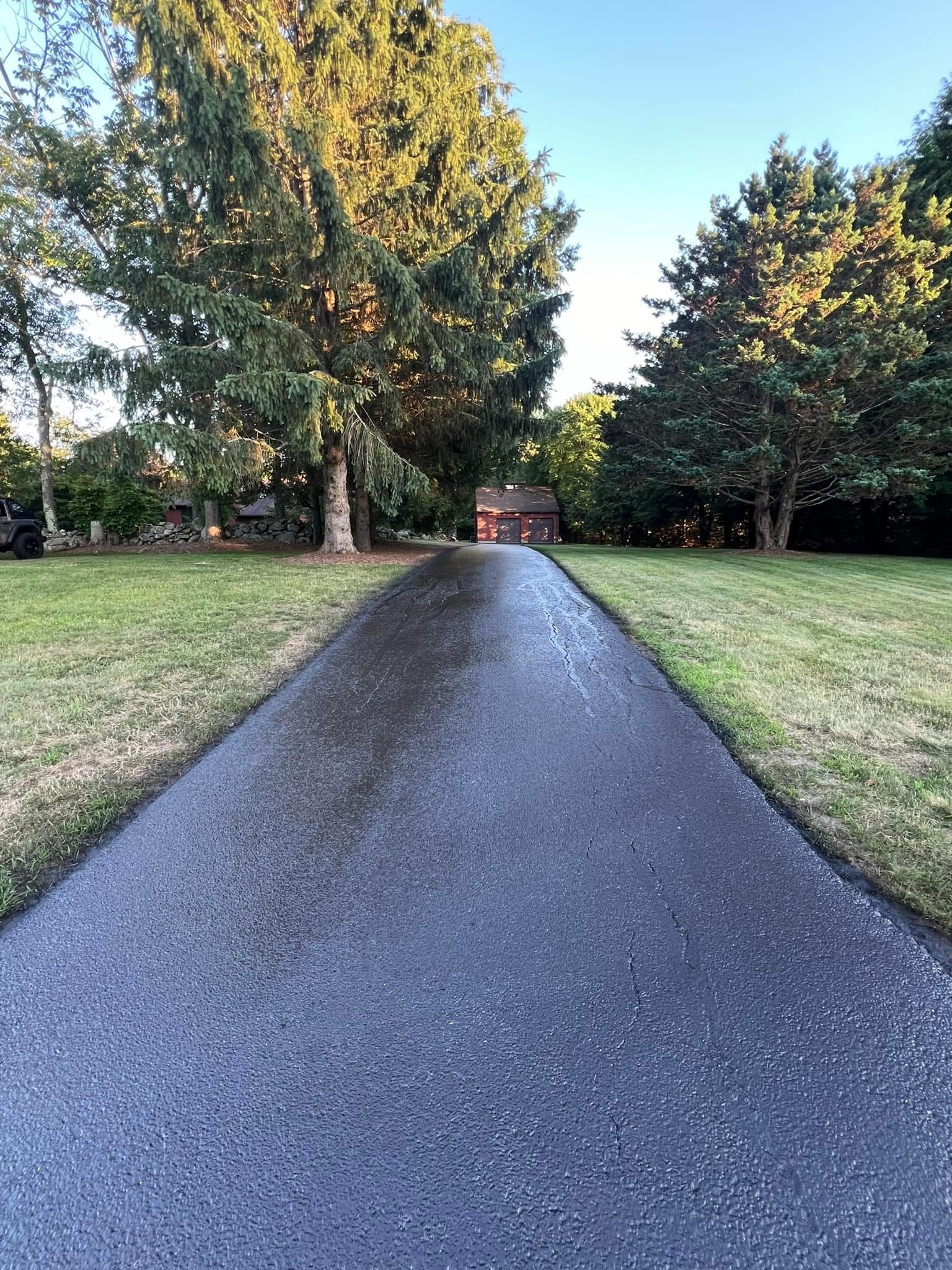 A dark asphalt driveway leads through a grassy yard toward a small brown building framed by two large, leafy trees.