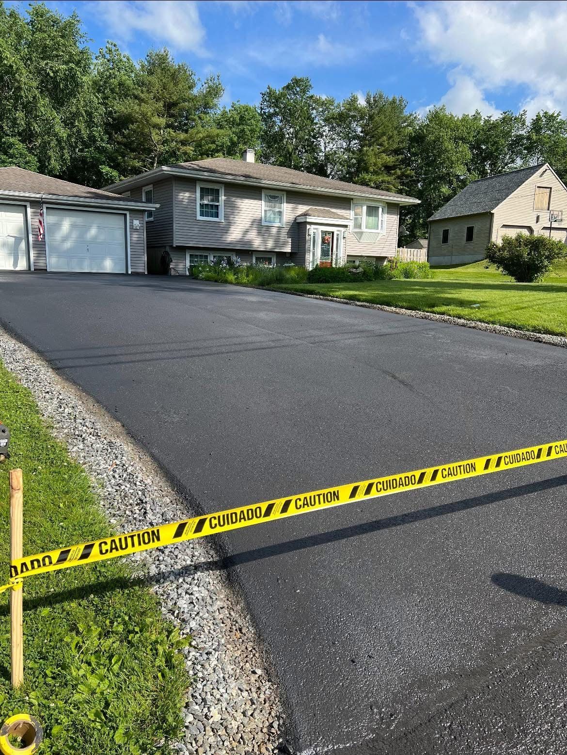 A newly paved dark asphalt driveway leads to a suburban house, cordoned off by yellow caution tape.