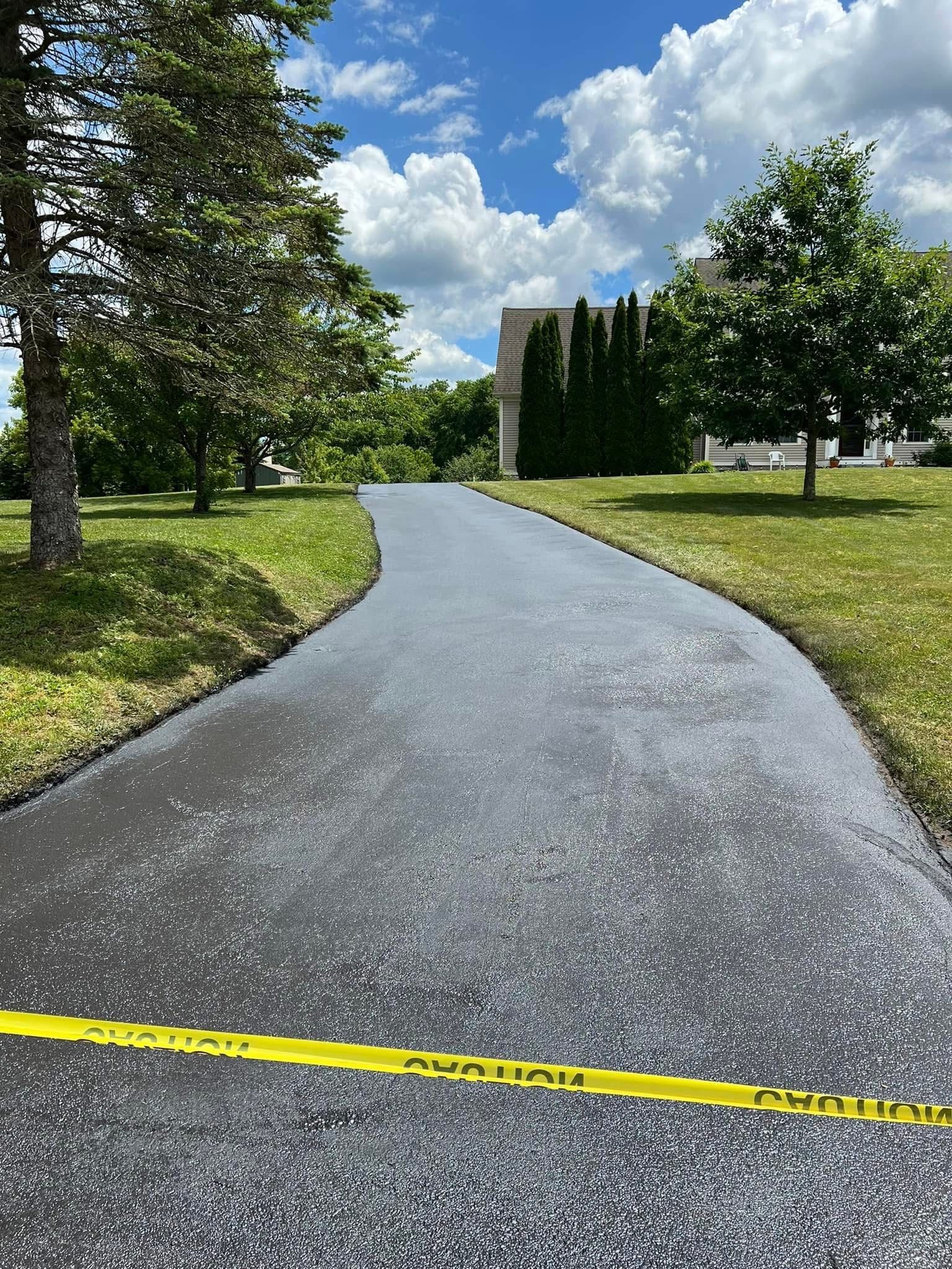 A newly paved asphalt path winds through a grassy park under a cloudy sky, blocked by a yellow caution tape.