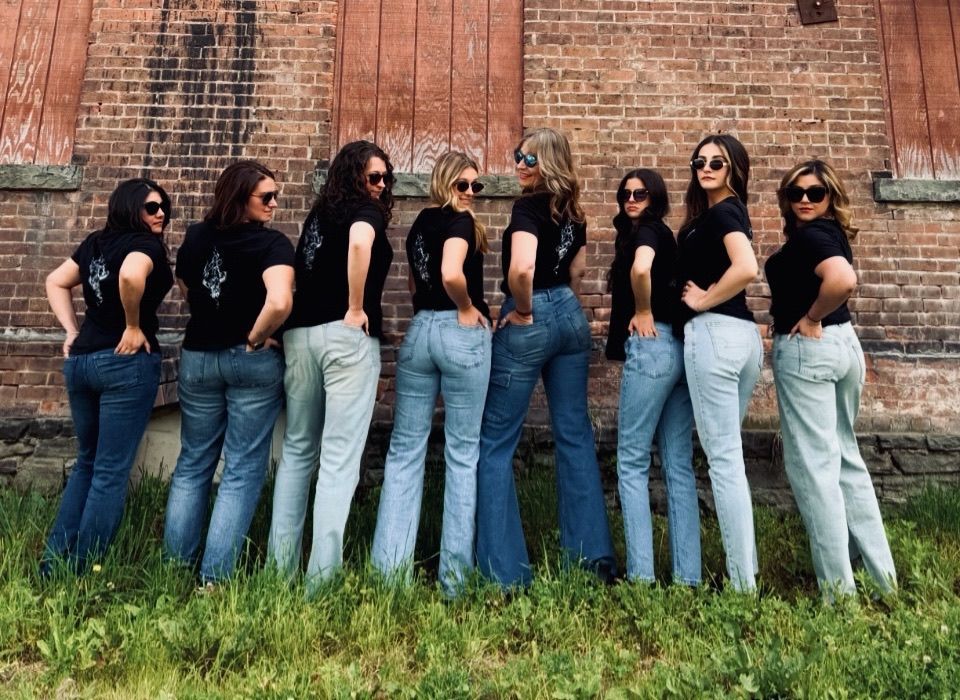 Eight people in black shirts and jeans standing in a line against a brick wall outdoors.