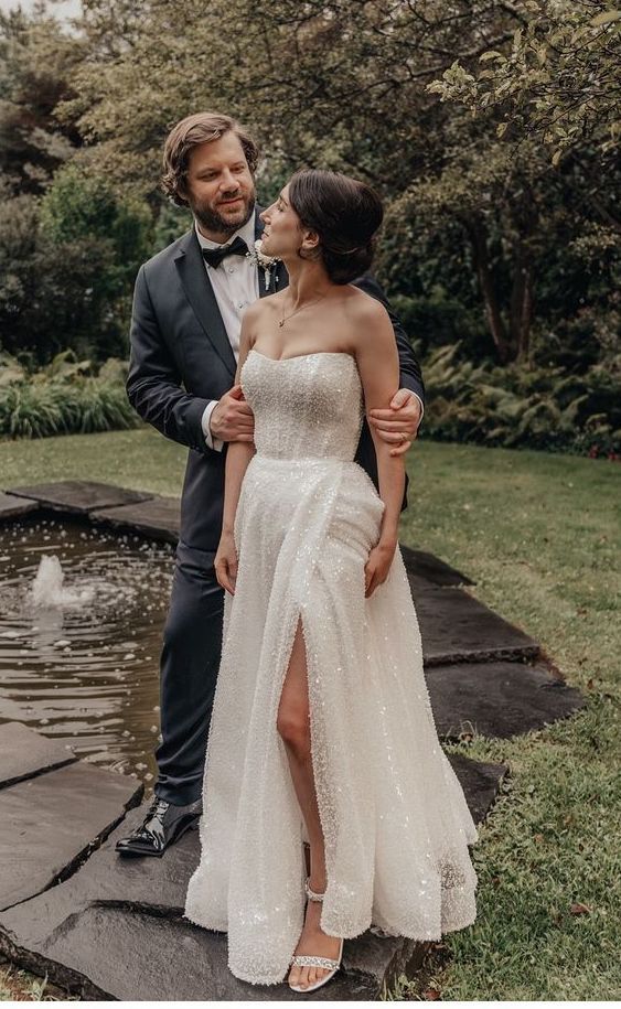 Couple stands near a water feature, woman in wedding dress with leg slit, man in suit.