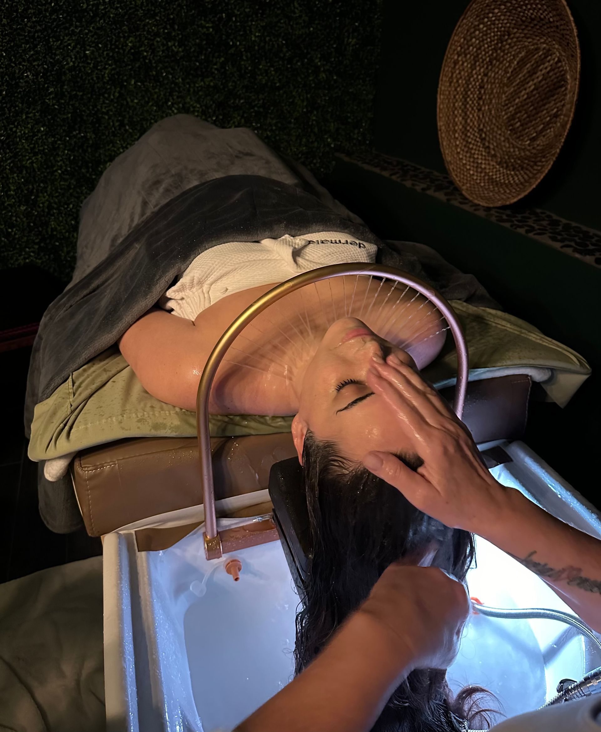 Woman having hair washed at a salon, face up. Water runs from a ring over her head, and someone washes her hair.