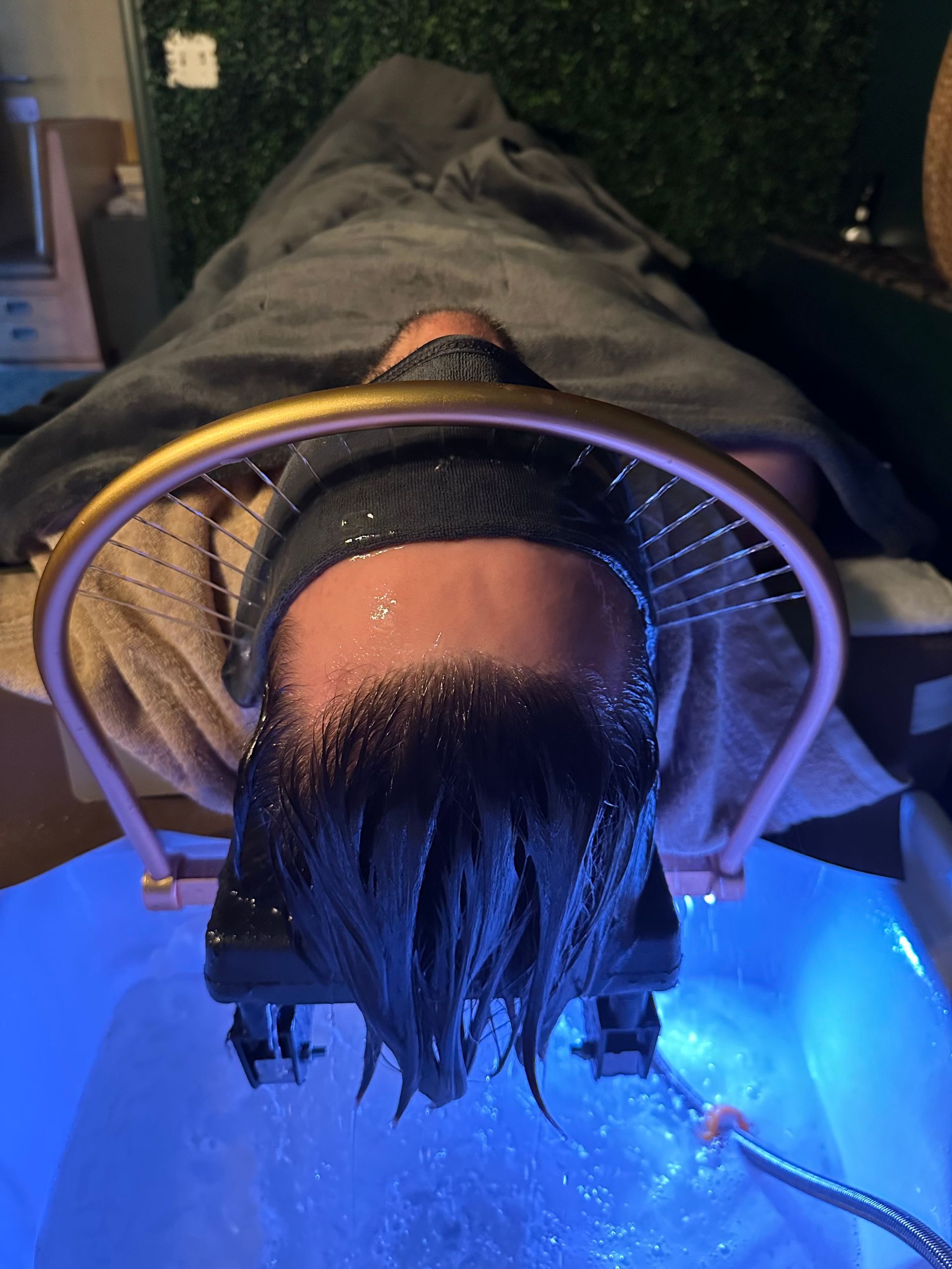 Person receiving a hair treatment; head over a basin with blue water. A gold shower arch is over the head.