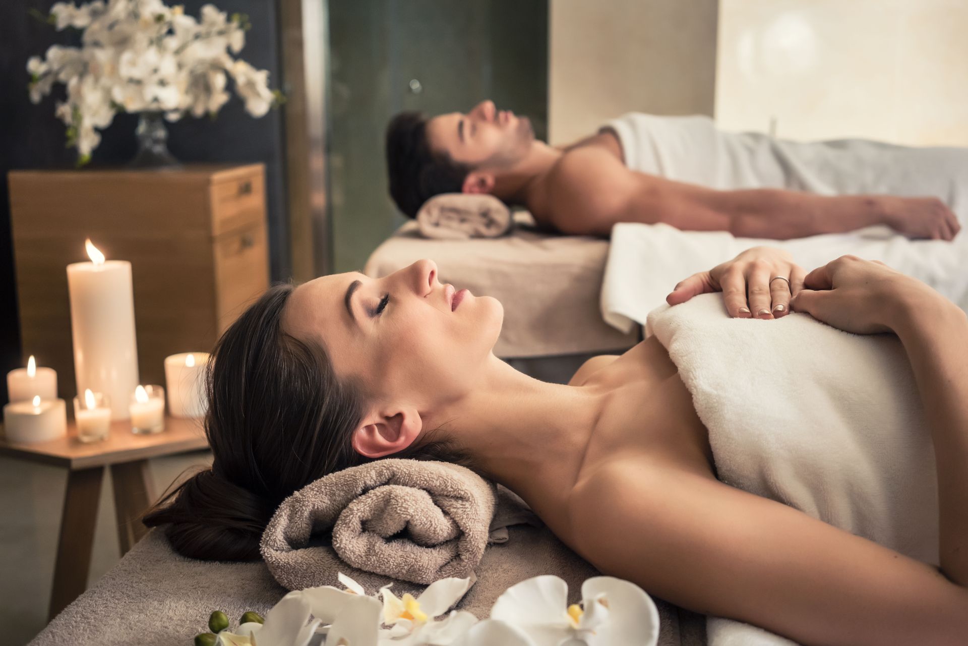 Couple receiving spa massages, relaxing with eyes closed on treatment tables, surrounded by candles.