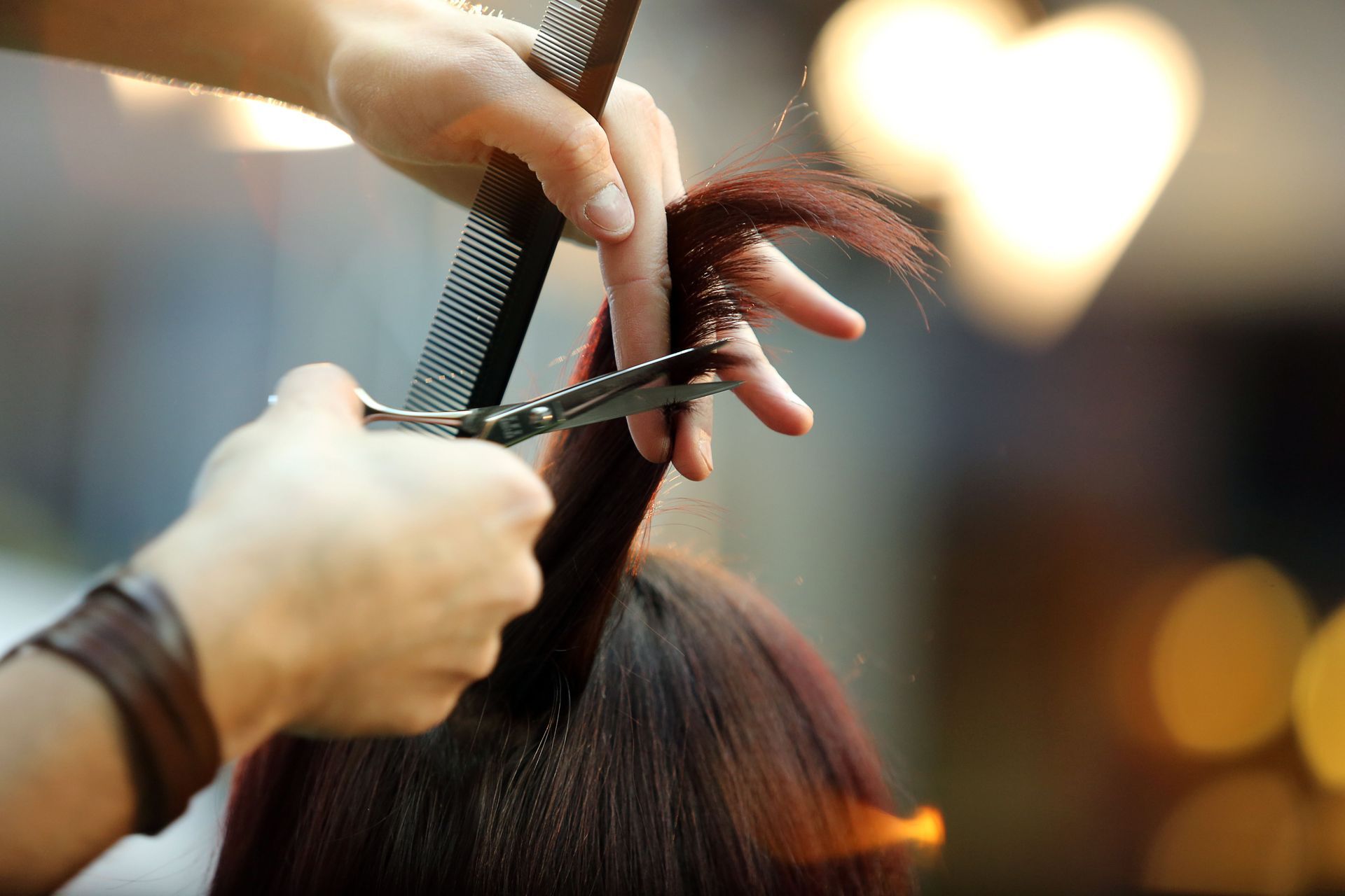 Hairdresser trimming a client’s dark hair with scissors and comb in a salon, warm blurred lights in background