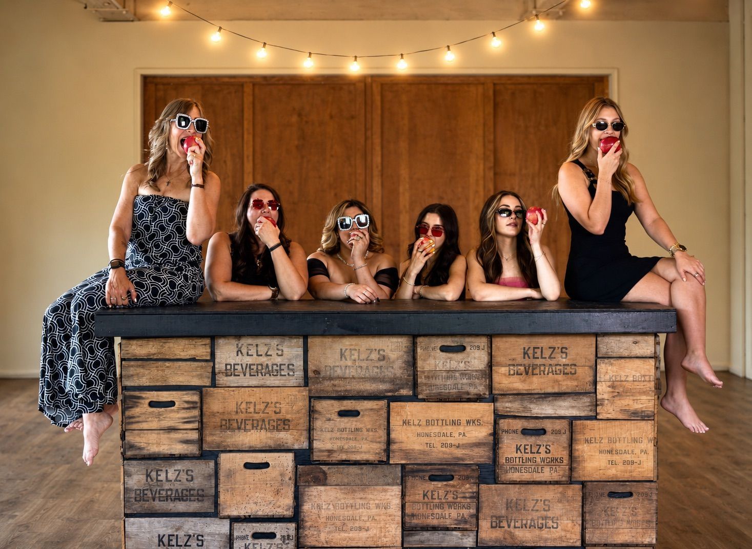Five women in sunglasses pose behind a rustic wooden bar decorated with string lights.