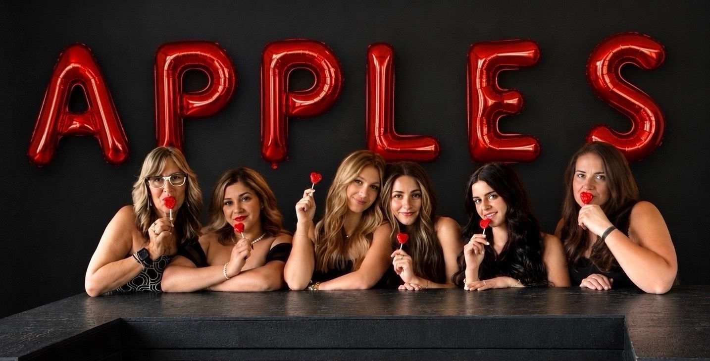 Three women posing behind a table with red balloons spelling “APPLES” against a black background
