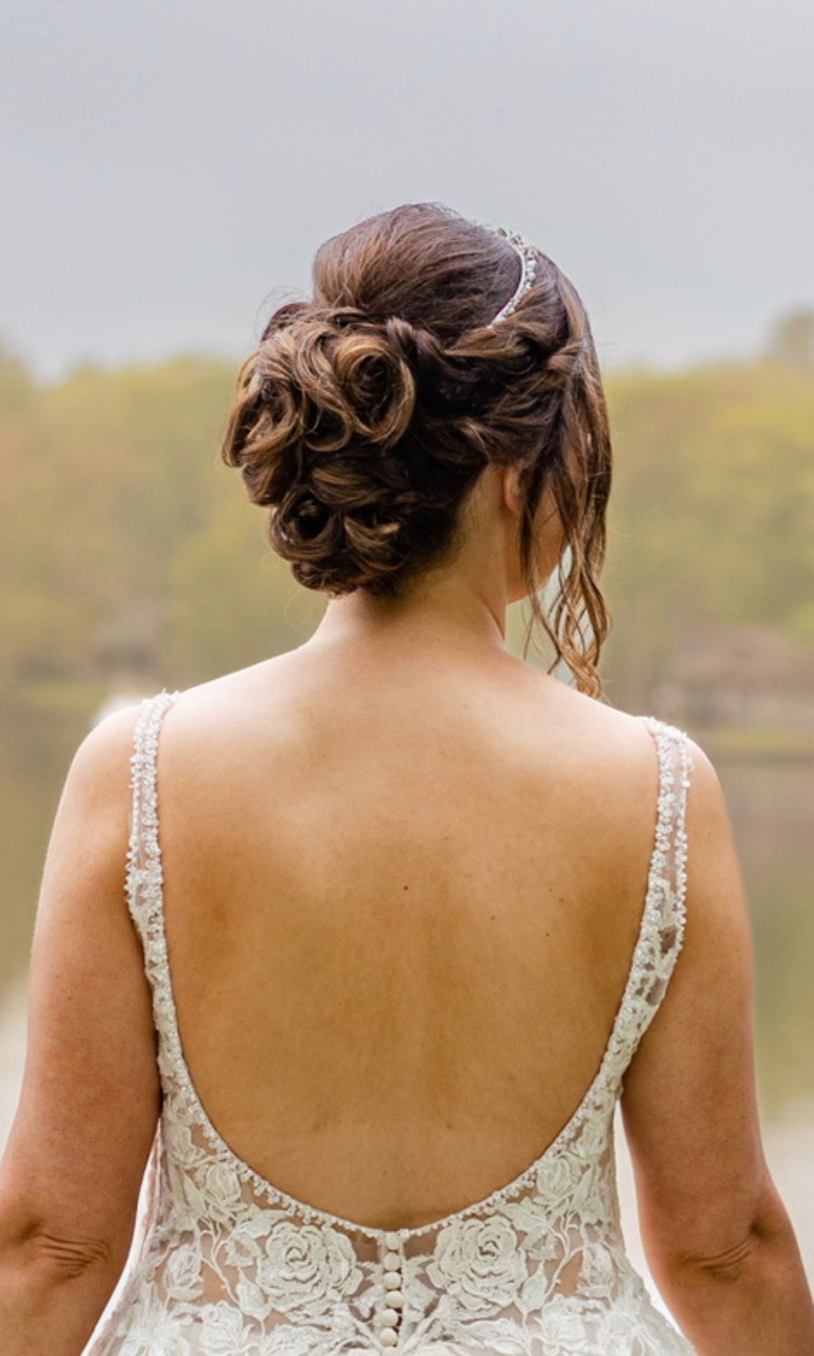 Woman from behind in a lace dress with an open back, hair in an updo, standing outdoors in a blurred natural setting