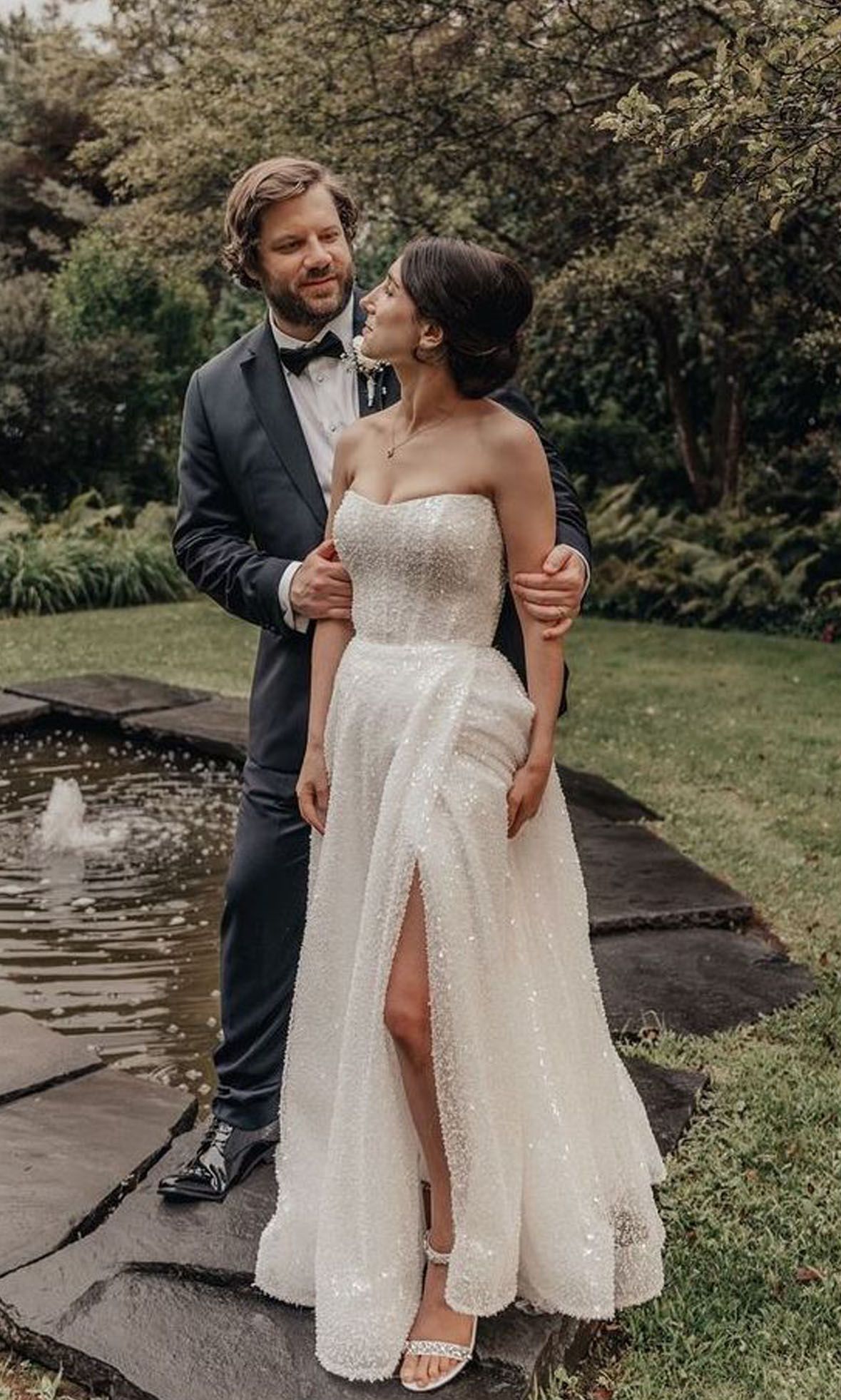 Couple in wedding attire standing by a garden pond, smiling at each other.