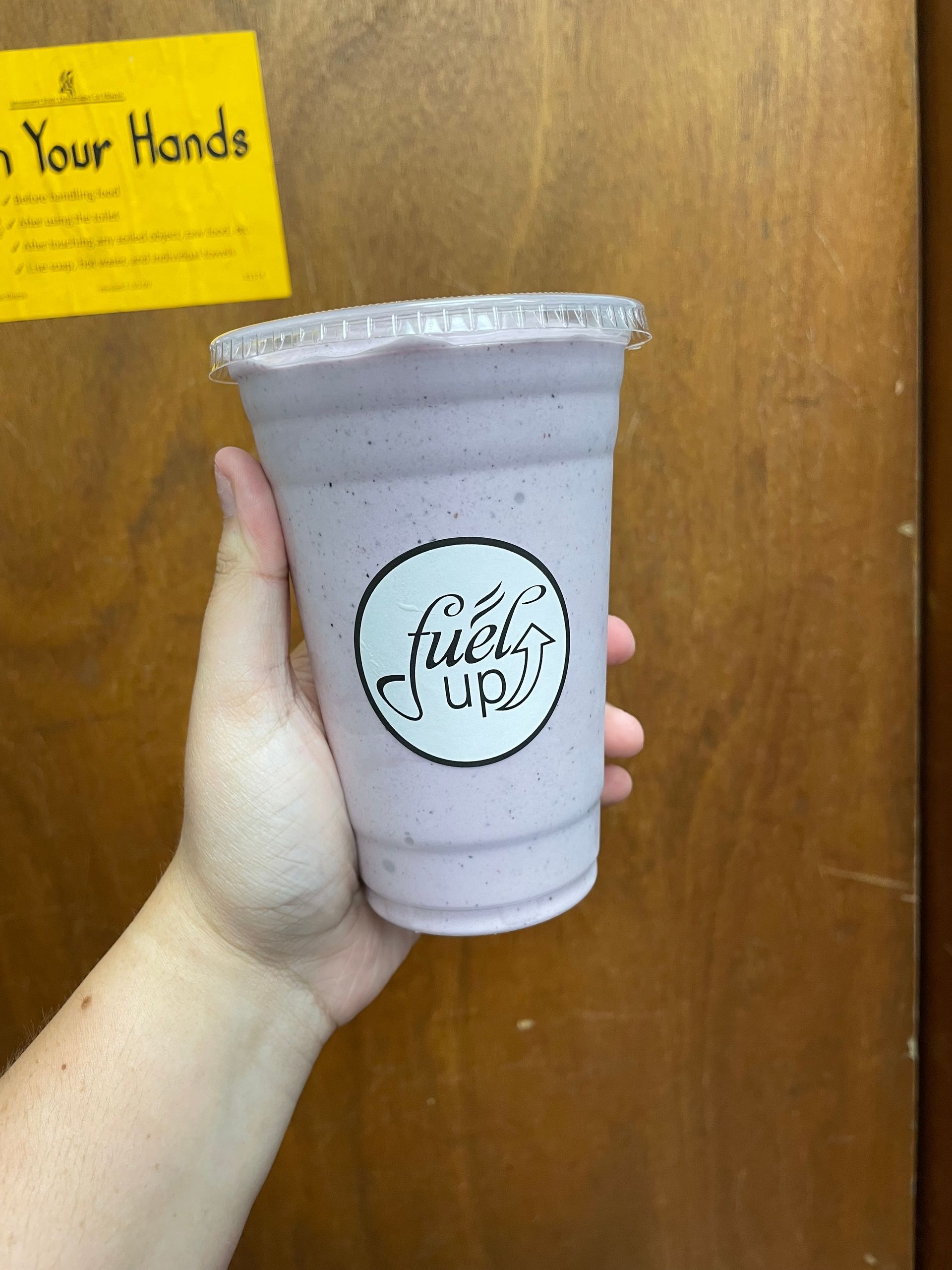 A person is holding a purple smoothie in front of a sign that says wash your hands