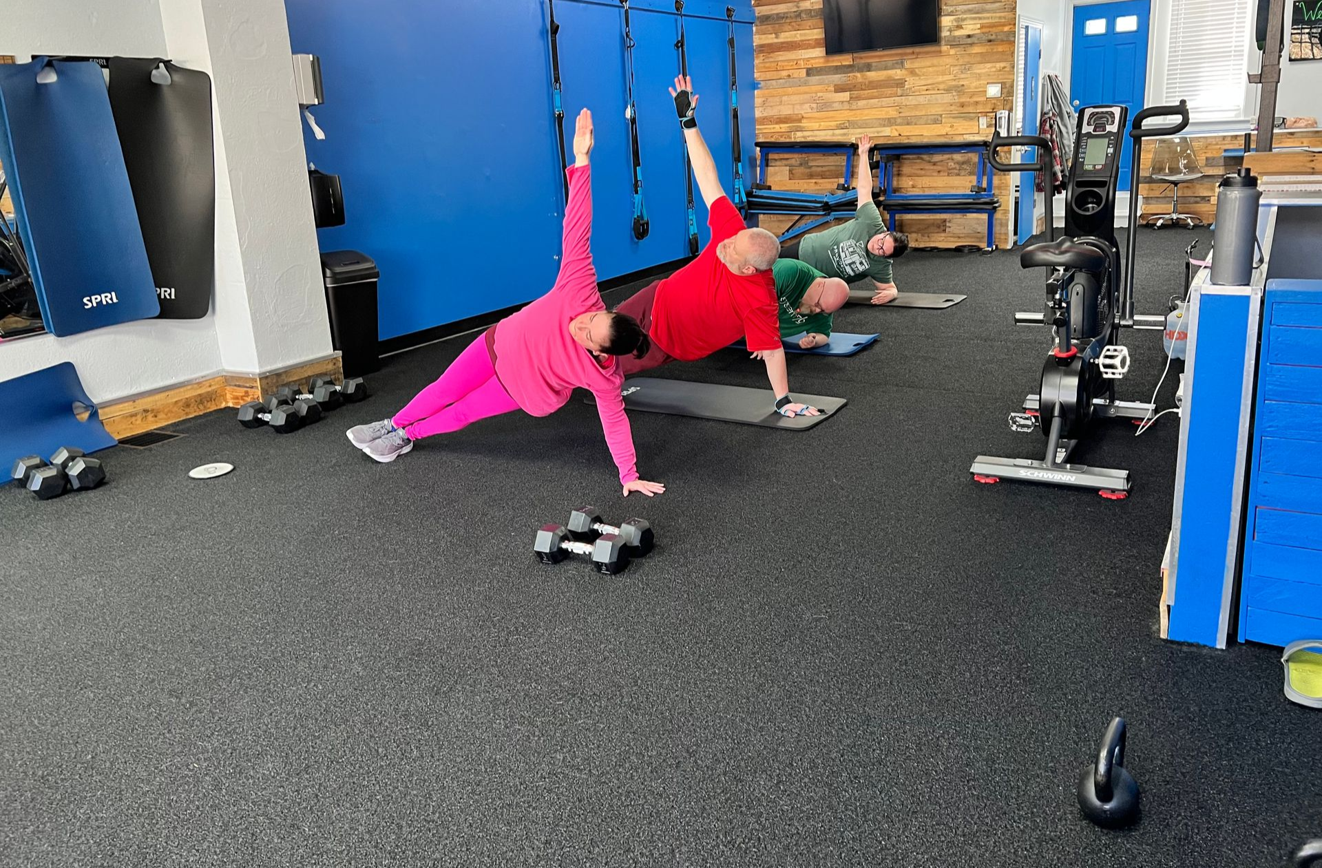 Three people doing side plank exercises in a gym with dumbbells. 