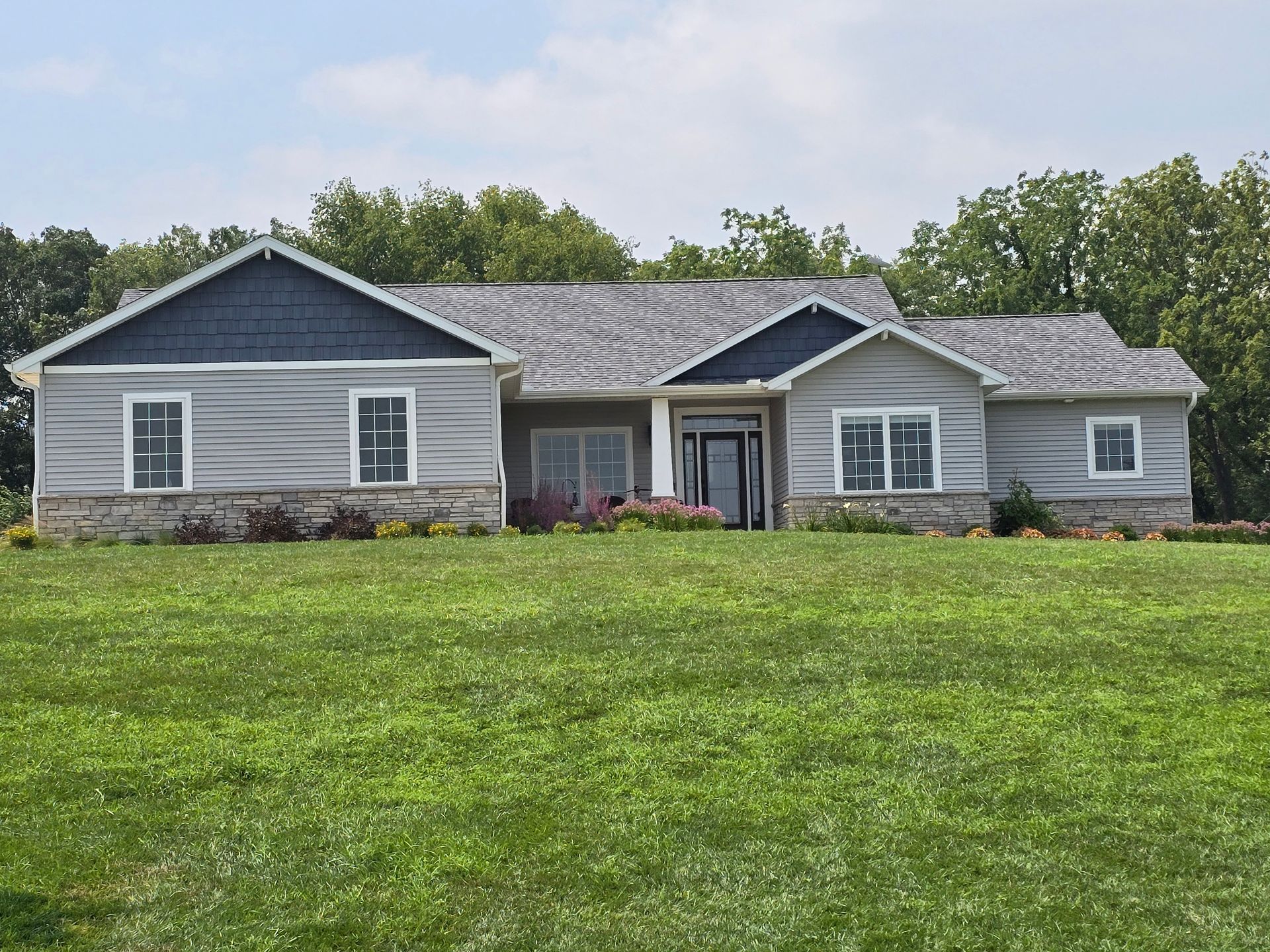 Ranch-style house with gray siding, stone accents, and blue roof against a green lawn and trees.