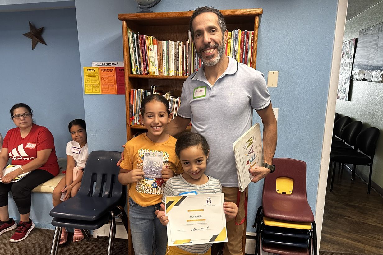 A man smiles with two children holding certificates near a bookshelf; others sit nearby.