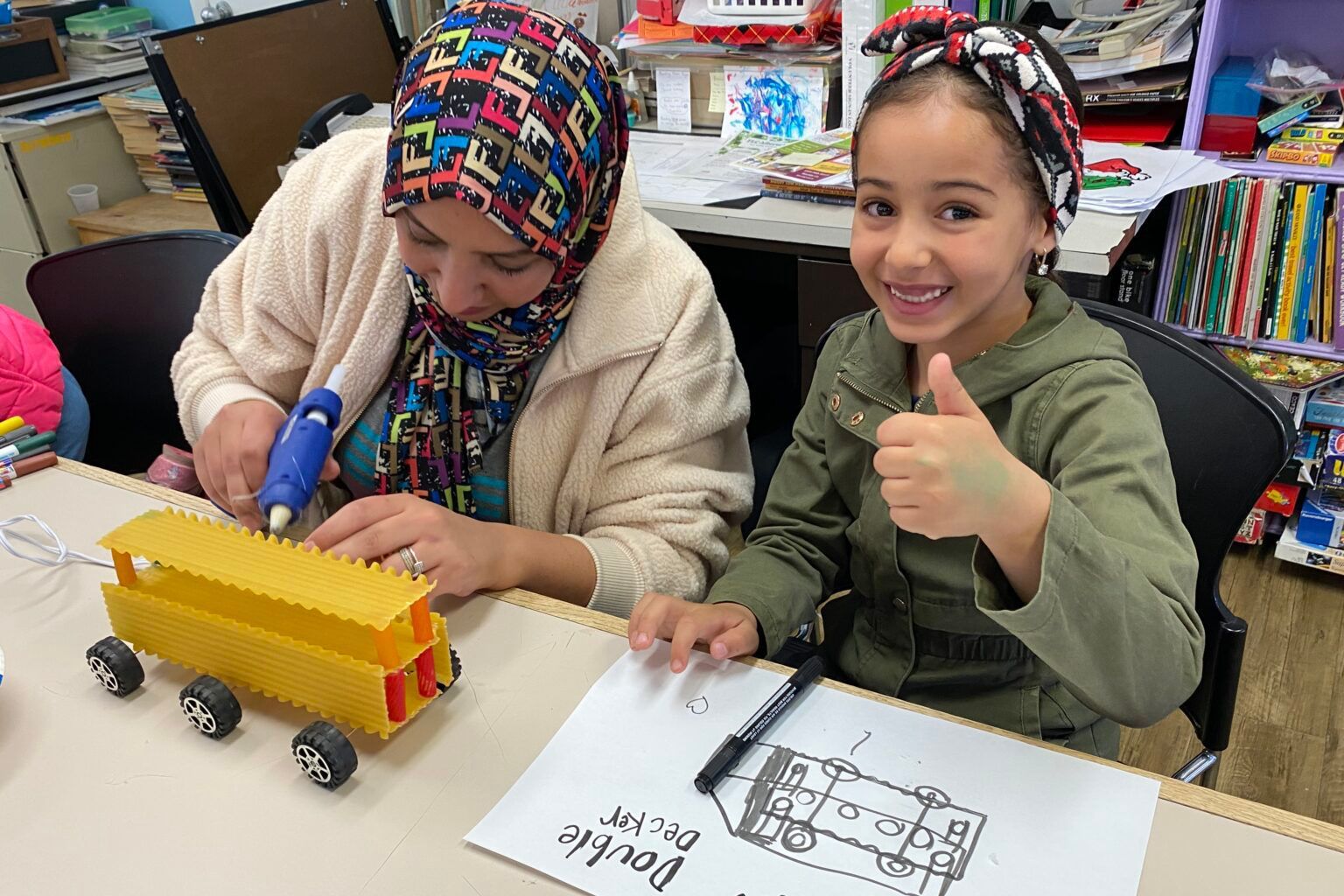 Woman and girl building a toy bus. The woman glues pieces while the girl smiles and gives a thumbs-up, with a drawing on the table.