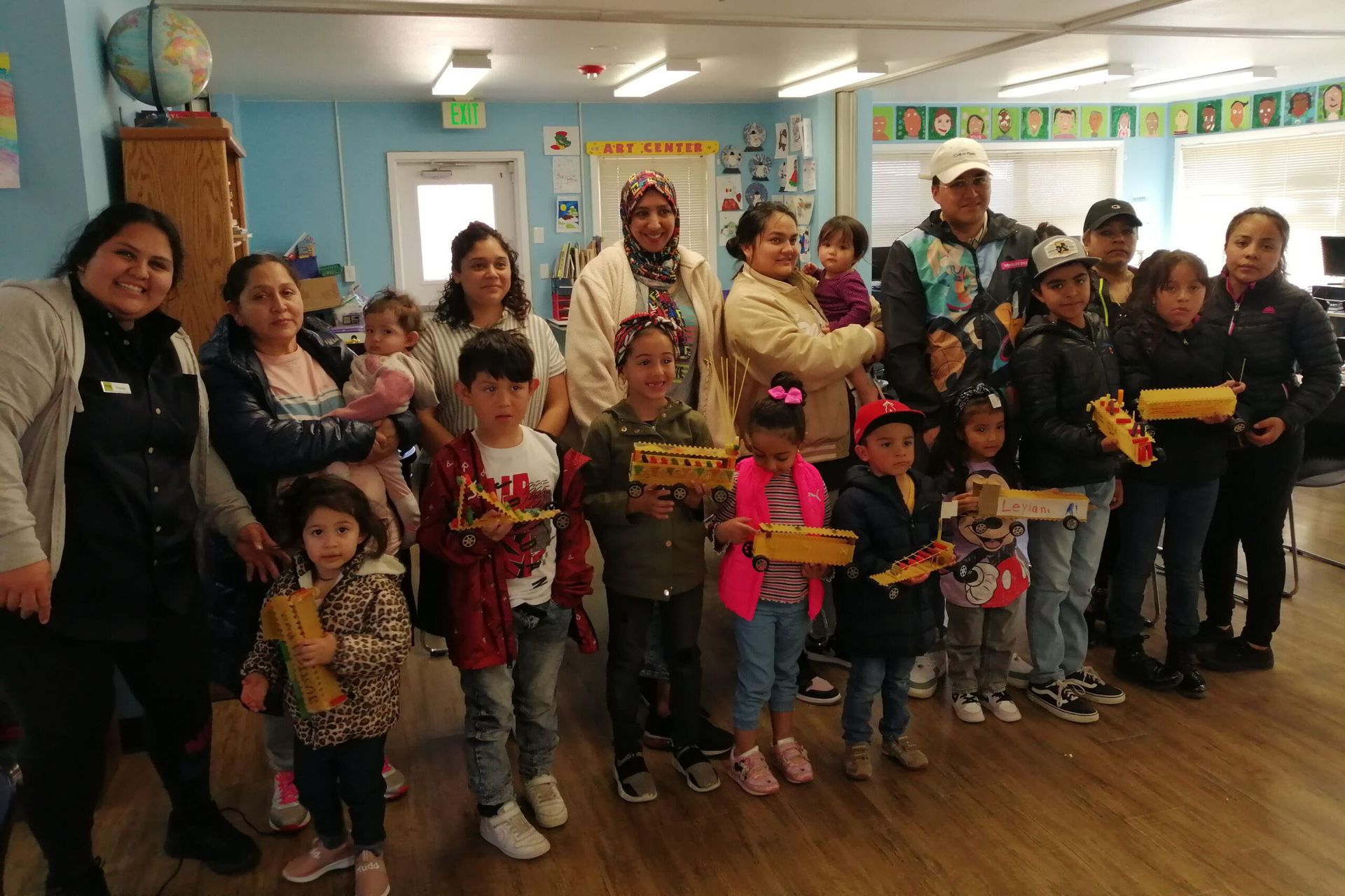 Group of adults and children holding toy trucks indoors. People of diverse ethnicities smile, likely at a community event.