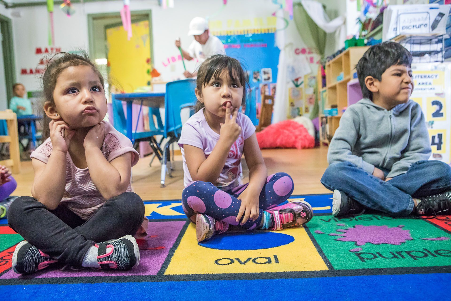 Three children sit cross-legged on a colorful rug in a classroom, looking thoughtful. A teacher stands in the background.