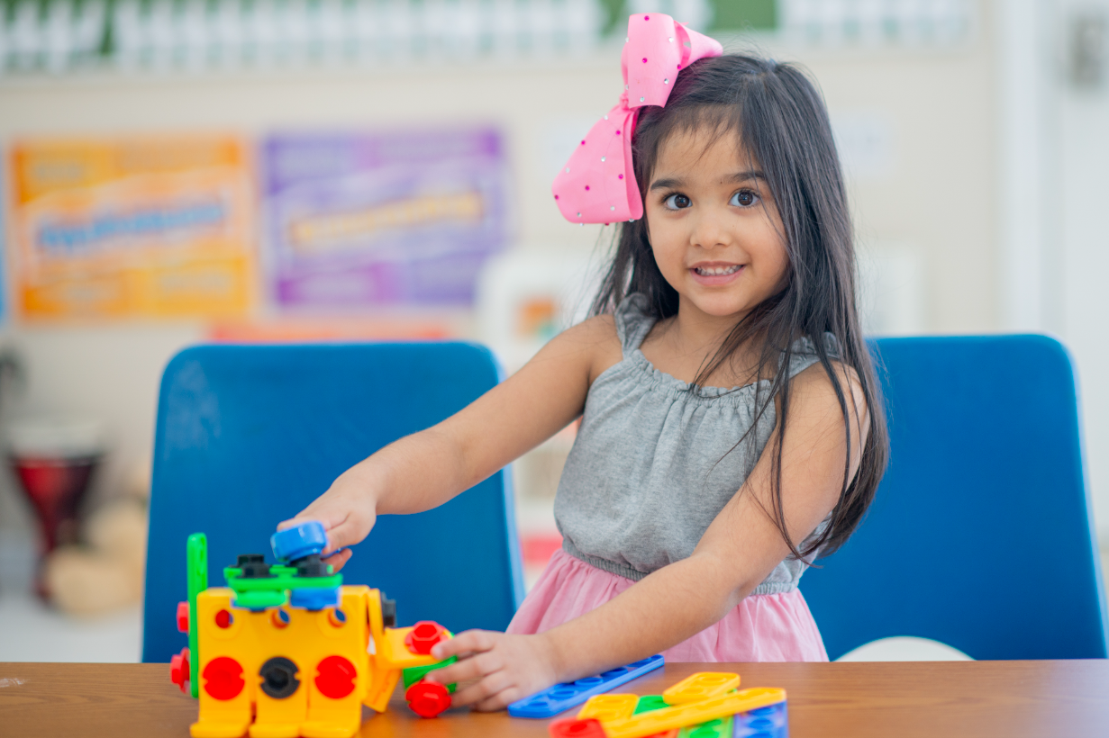 Girl with a pink bow plays with colorful blocks at a table, smiling in a classroom.