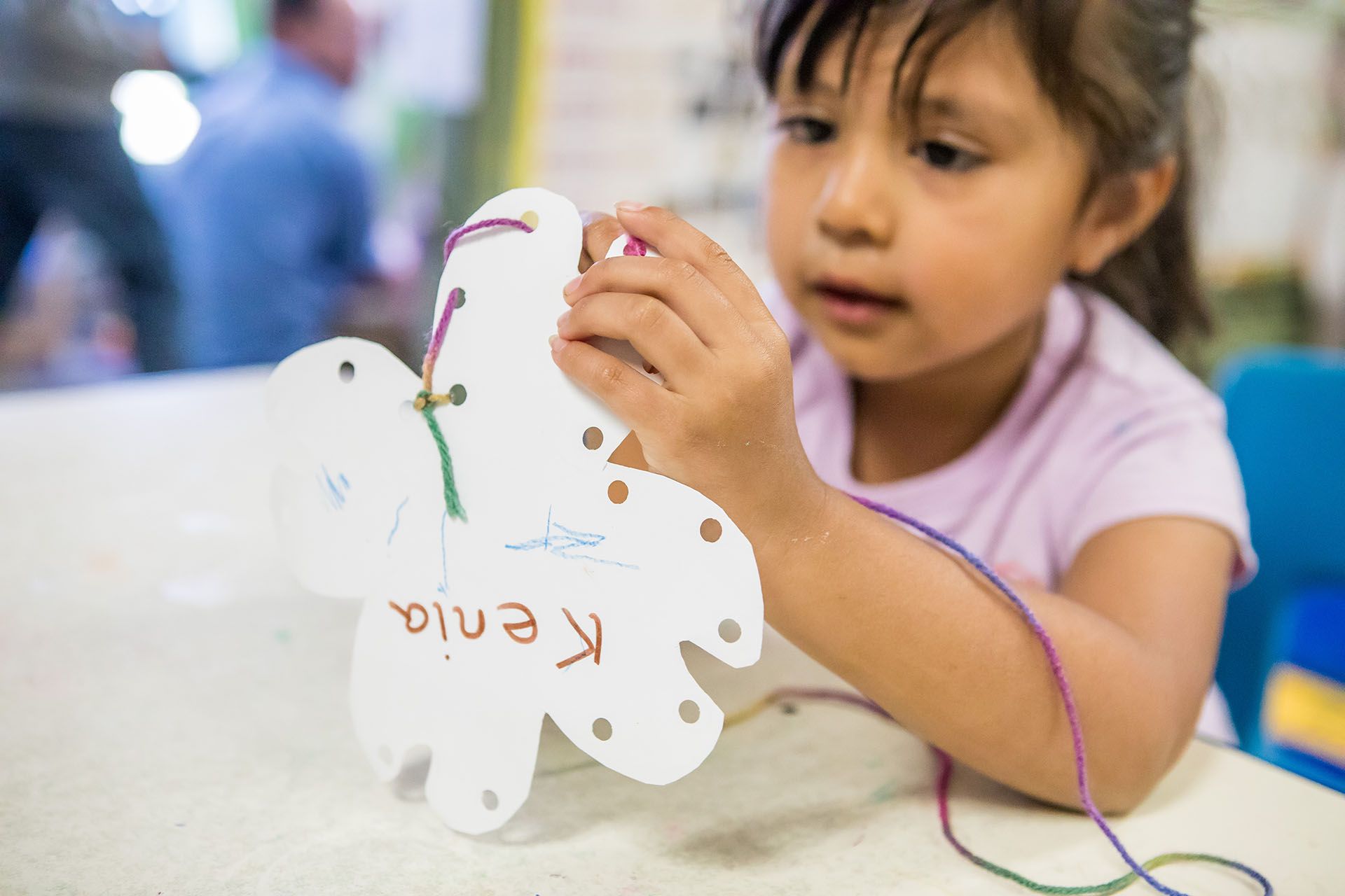 A young girl, Kenia, sews colorful thread onto a white paper cutout at a table in a classroom.