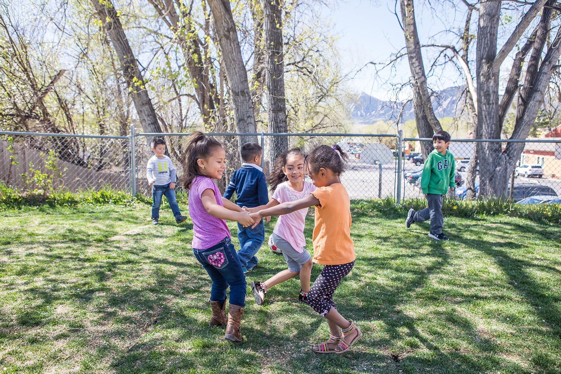 Children playing in a grassy yard, holding hands and smiling. Trees and a chain-link fence are in the background.