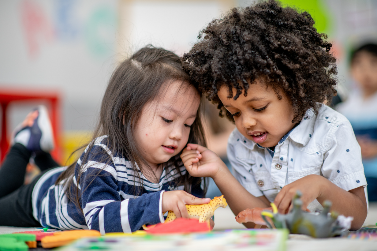 Two young children, diverse in ethnicity, playing together with toys on the floor of a classroom.