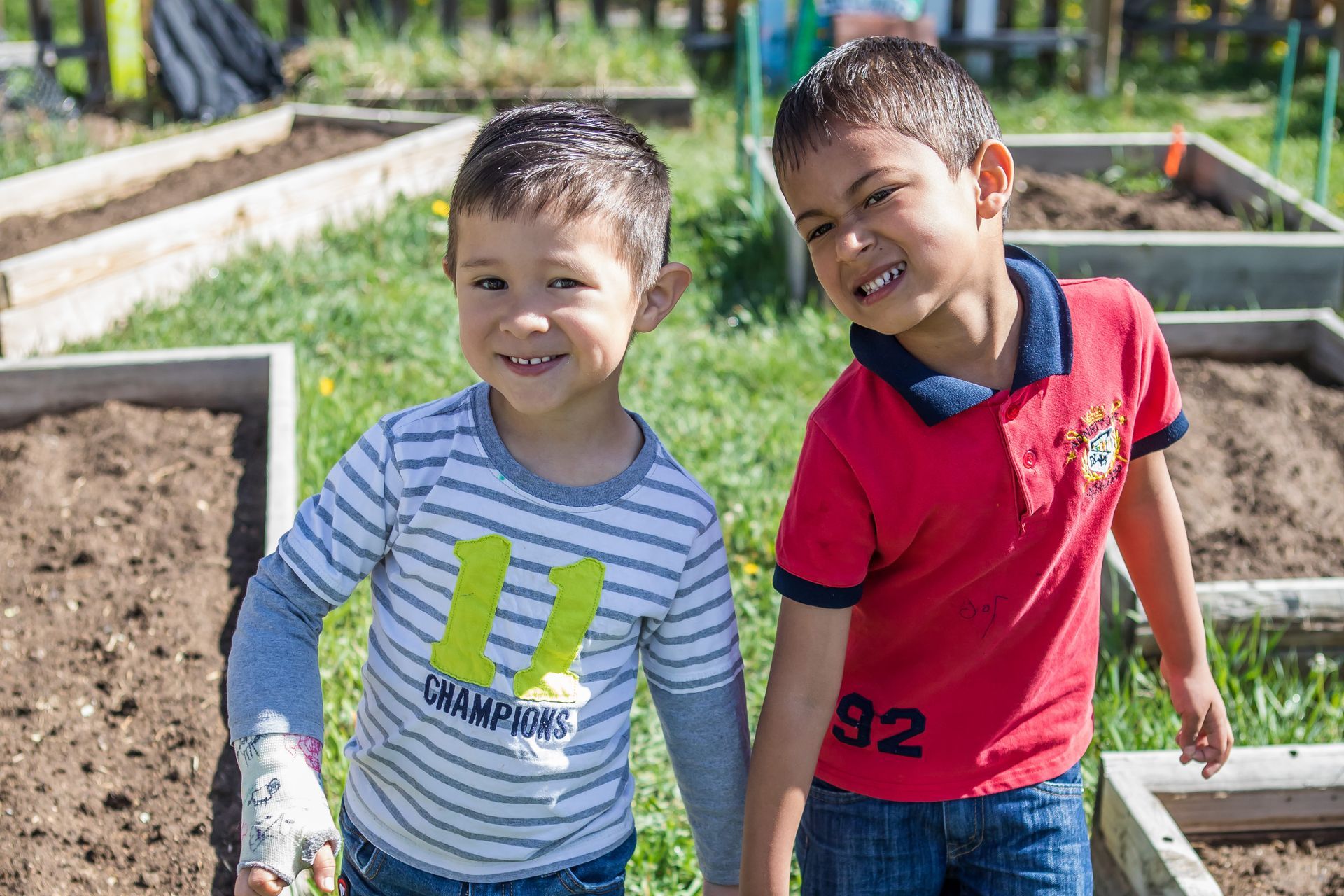 Two young boys holding hands smile in a garden with raised beds; one wears a striped shirt, the other a red polo.