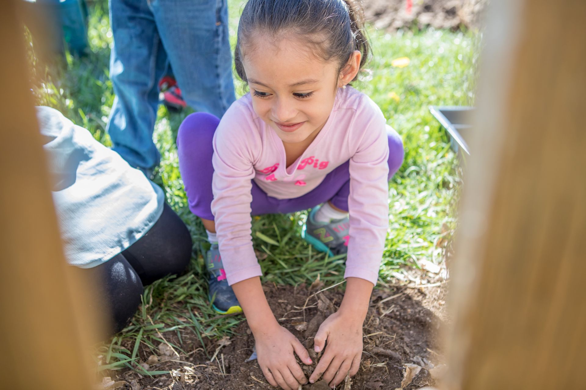 Young girl in pink shirt and purple leggings squats, planting in a garden, with other people nearby.
